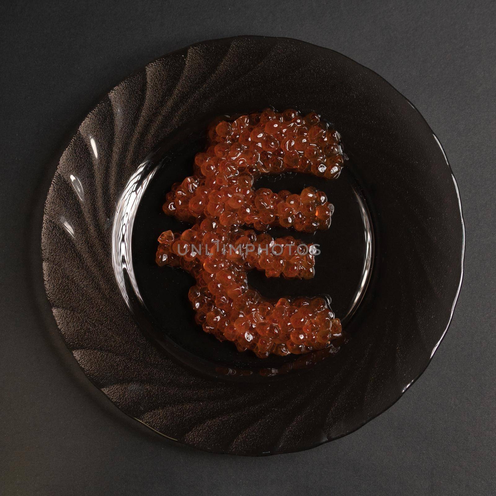The symbol of the euro is beautifully assembled with real red caviar on a black plate on a gray background view from above. Food in the world's elite restaurants is a symbol of money