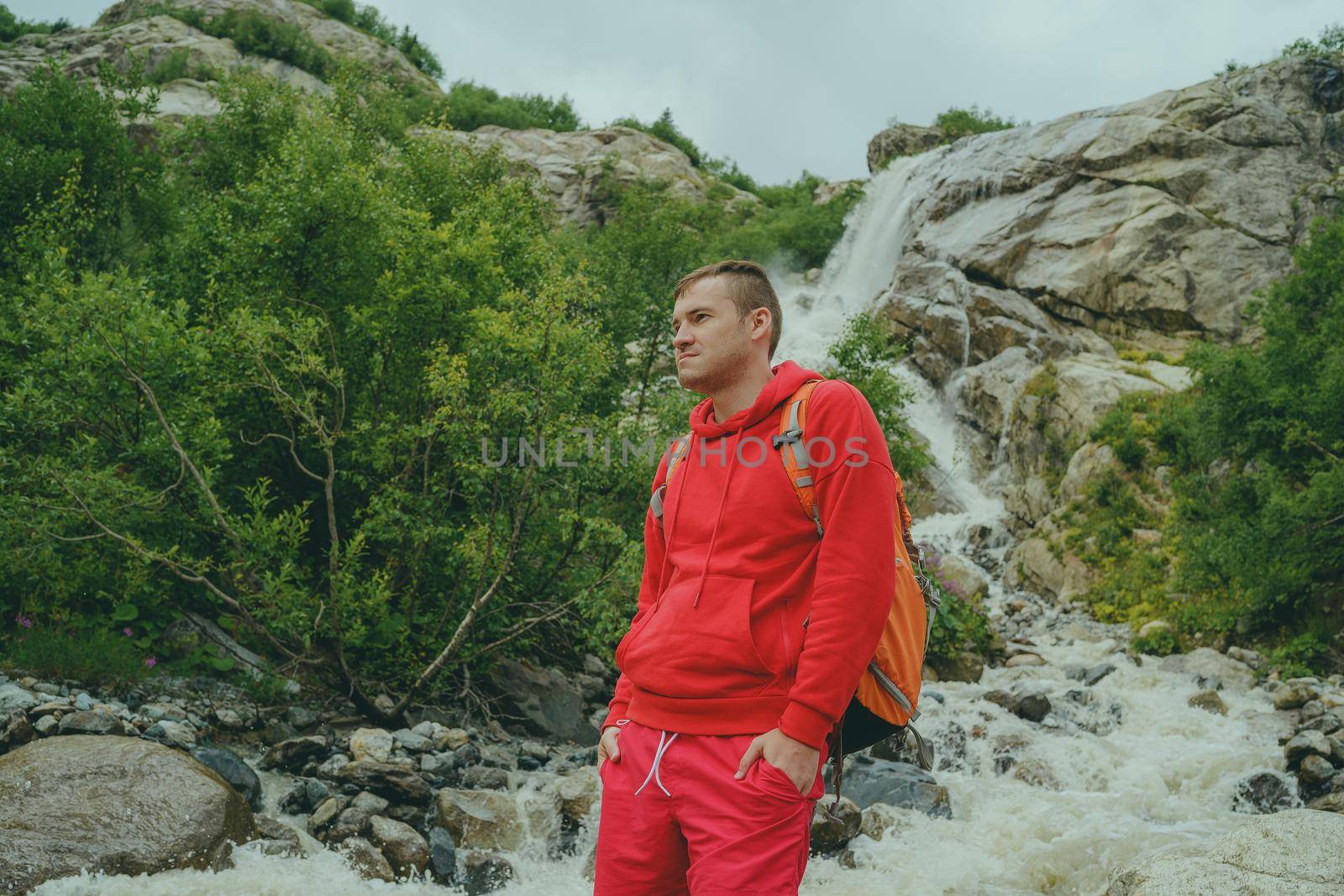 Portrait of young man looking away on background of waterfall. Adult male tourist enjoying beautiful view in mountainous area. by epidemiks