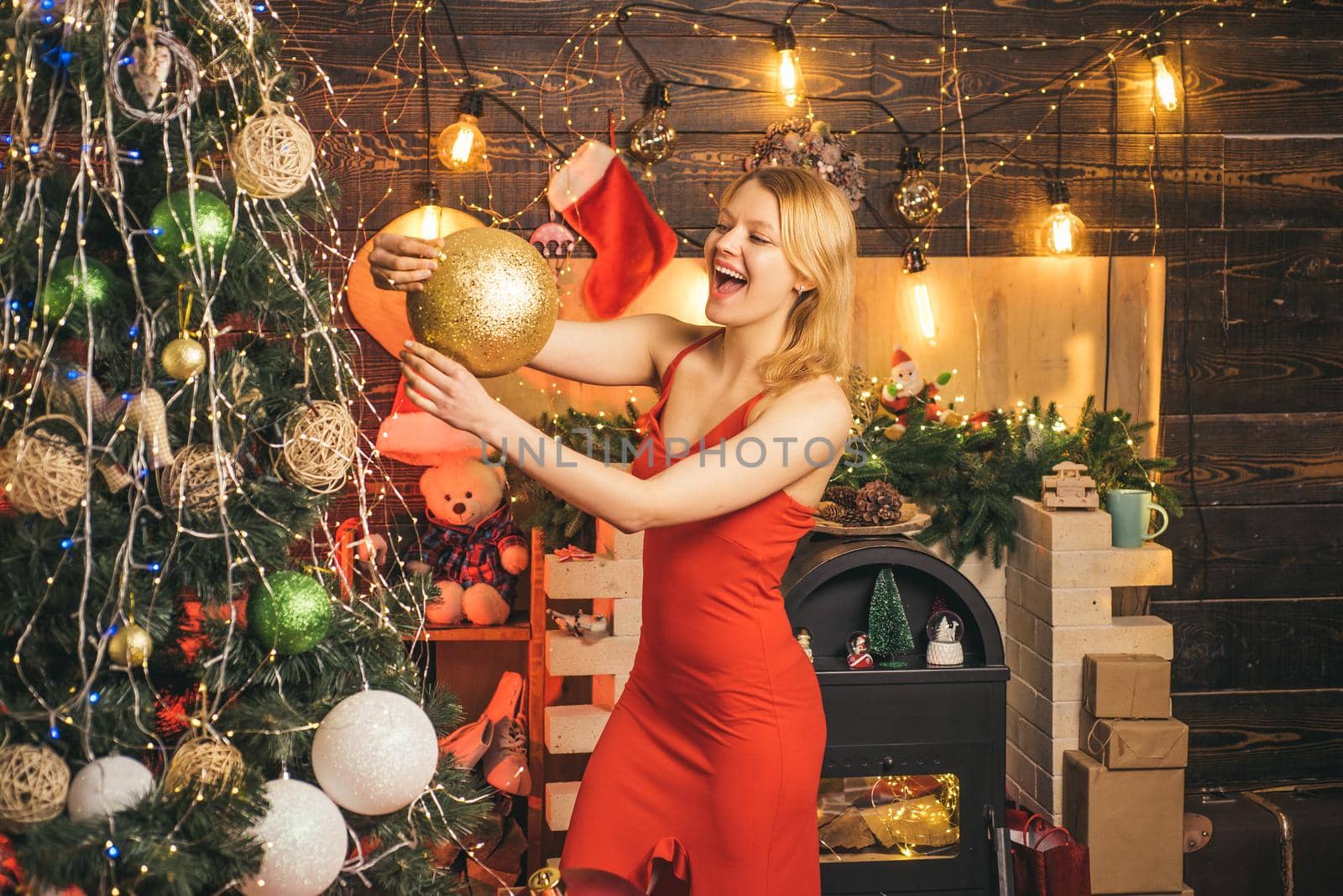 Cheerful smiling blonde woman in red dress decorates Christmas tree with gold ball. Christmas time. Christmas shopping concept. Happy holiday. Xmas tree. New Year surprise present