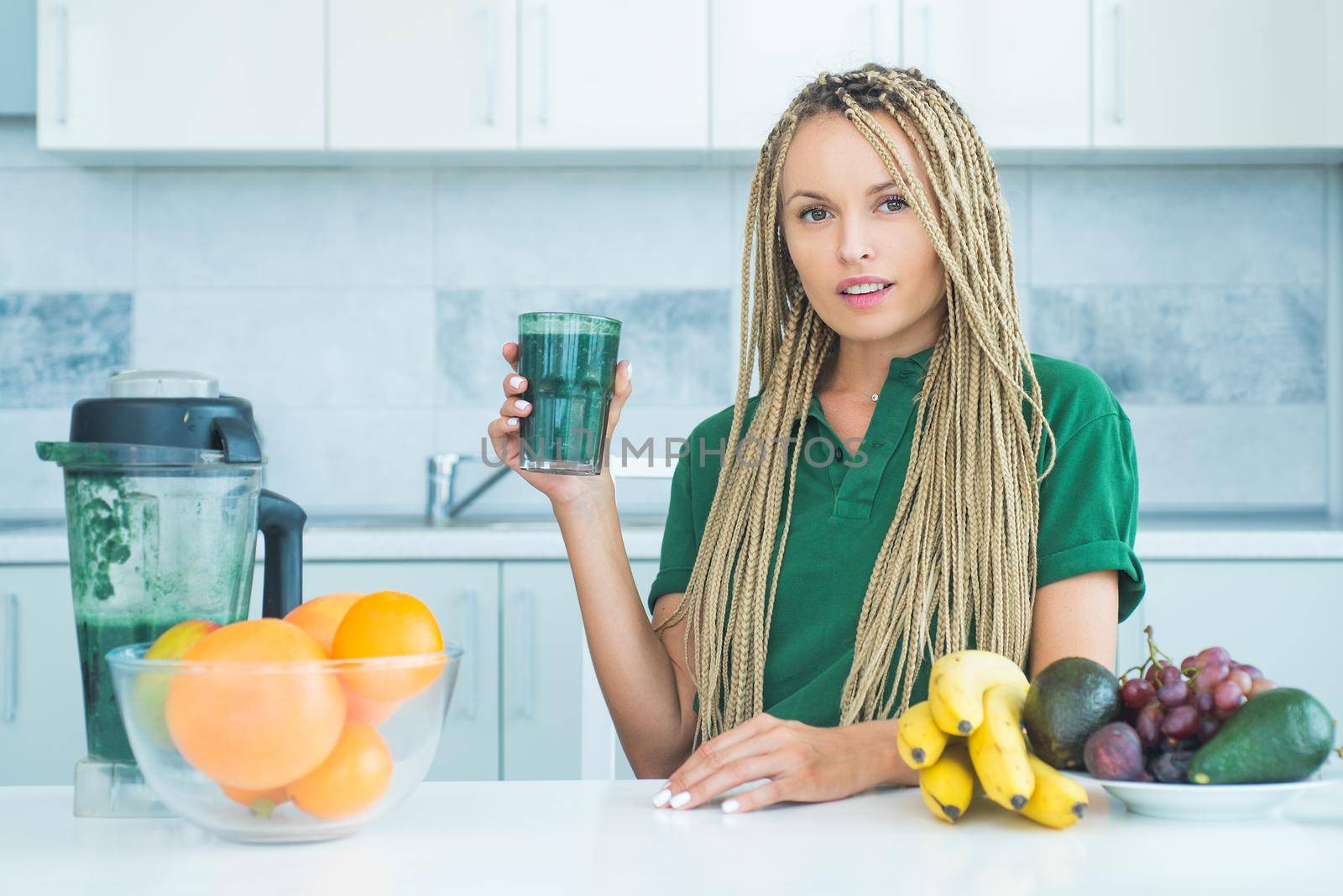 Positive woman hold cup of fresh green smoothie. Eco lifestyle. Young woman drinking green smoothie in the kitchen with fruits and vegetables. Vegan meal and detox menu