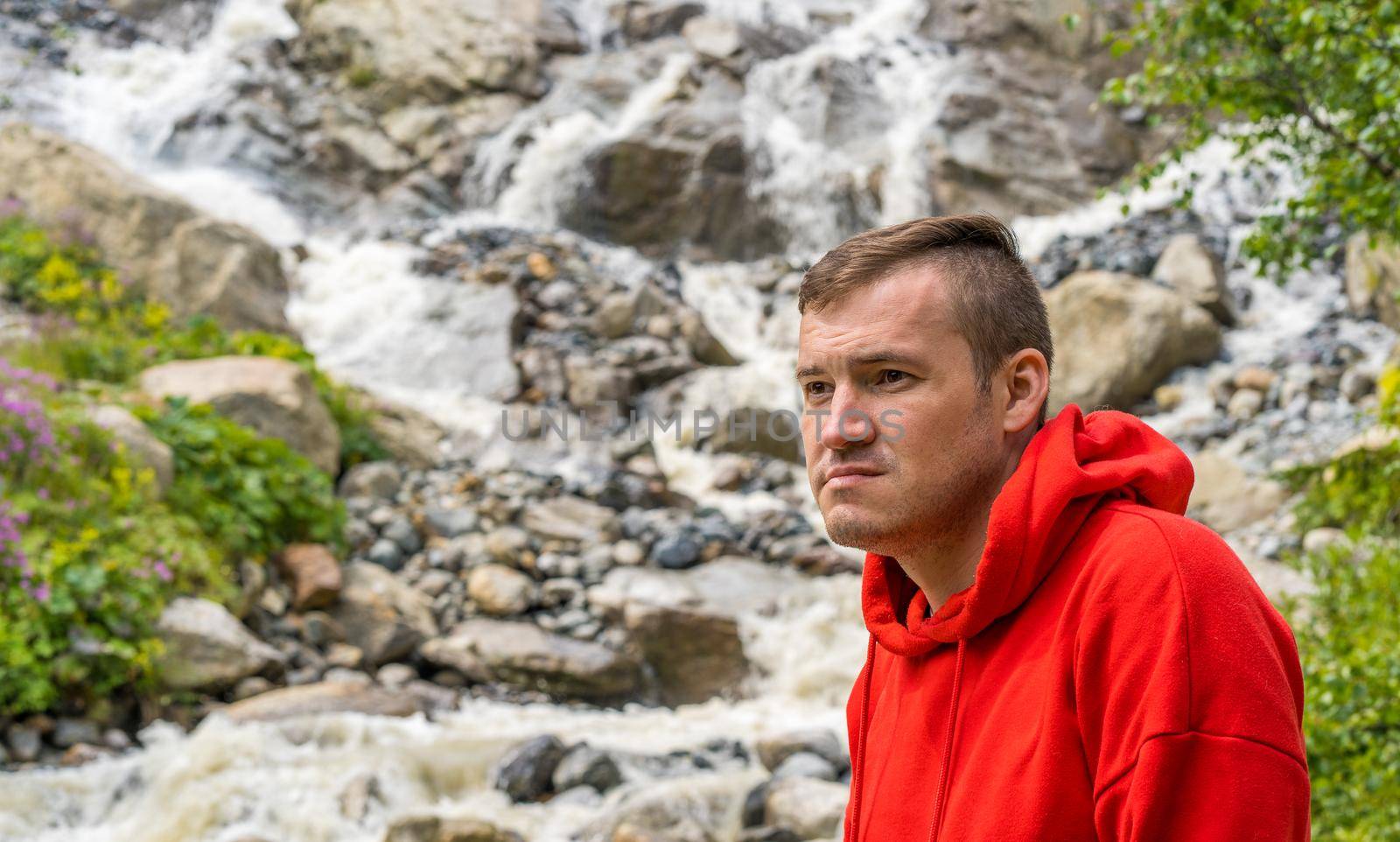 Portrait of young man looking away on background of waterfall. Adult male tourist enjoying beautiful view in mountainous area. by epidemiks