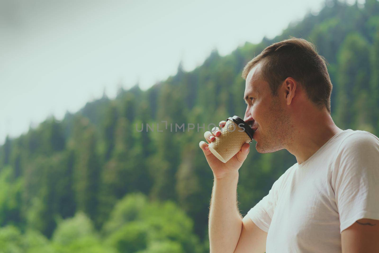 Portrait of pleasant guy spending time enjoying vacation and drinking coffee on background of mountain forest. Adult male enjoying hot drink against mountain landscape. by epidemiks