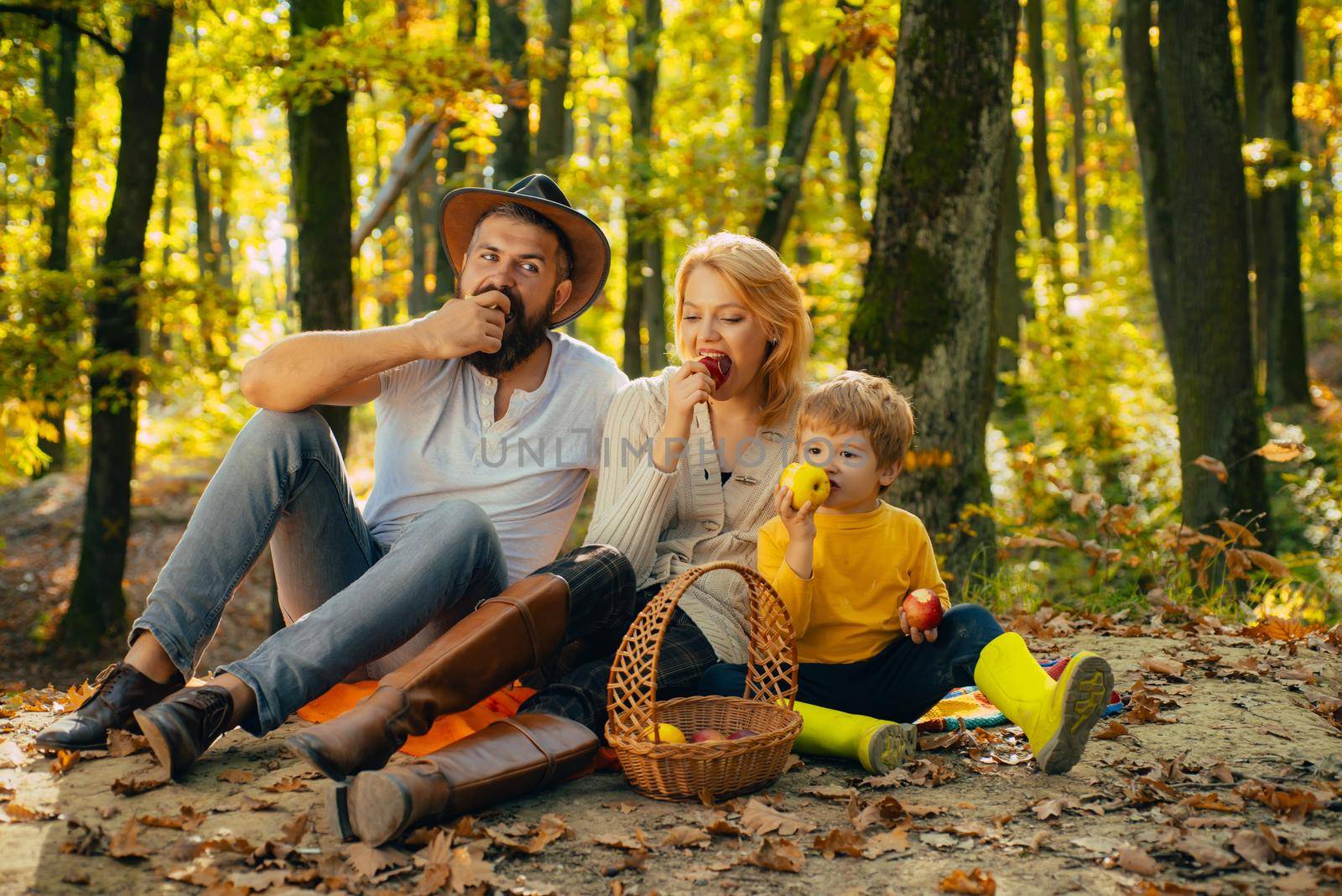 Cheerful family sitting on the grass during a picnic in a park. Mom, dad and baby happy relax at sunset. There is a basket with meal and toys for the kid. Autumnal family walk concept. by Tverdokhlib