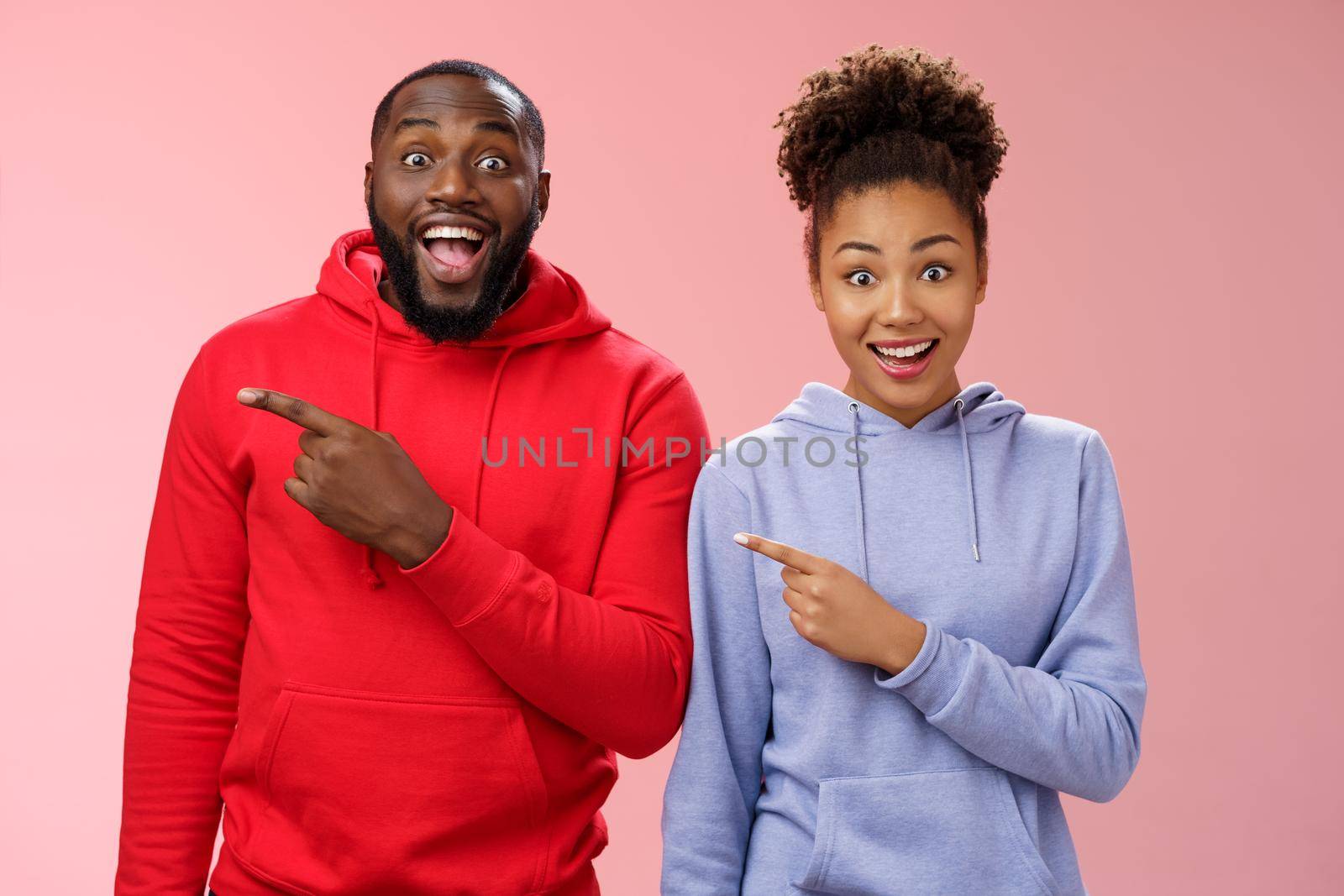 Impressed two best friends african american man woman 25s drop jaw amused visit awesome interesting park pointing left index fingers widen eyes thrilled having fun together, standing pink background.