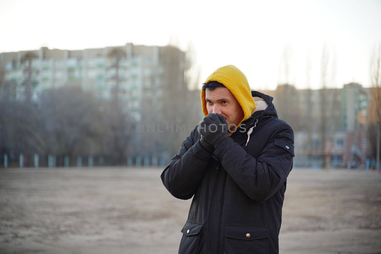 Young frozen man in yellow hood and black jacket. Adult male warming hands in gloves with his breath in cold weather