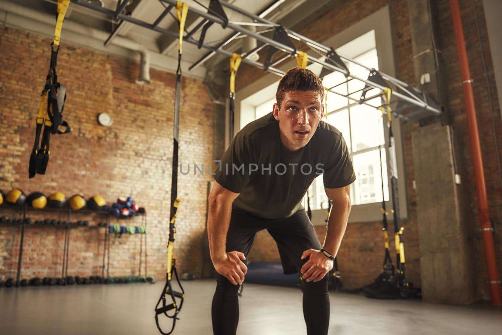 Time to rest. Young athletic man in sportswear looking exhausted after workout while standing at loft style gym. Tiredness. Professional sport. TRX Training.