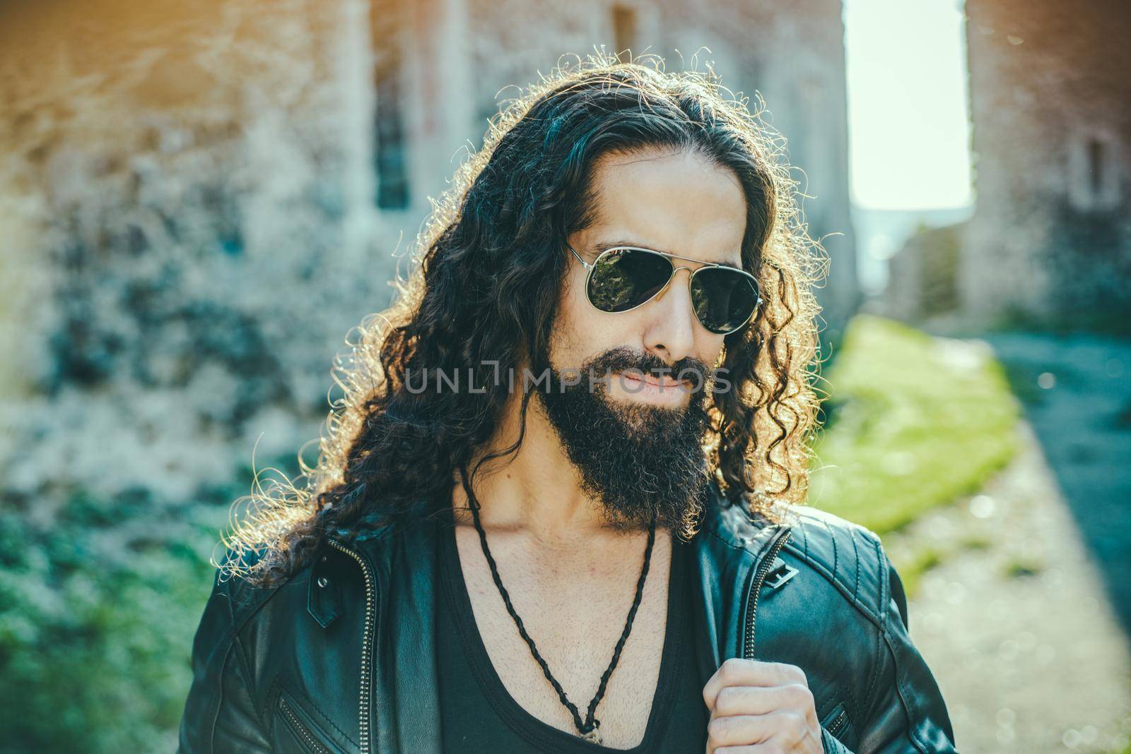 Self confident handsome man in sunglasses. Macho man walking outside alone. Stylish young fashionable man with long wavy hair wearing bikers leather jacket and aviator sunglasses