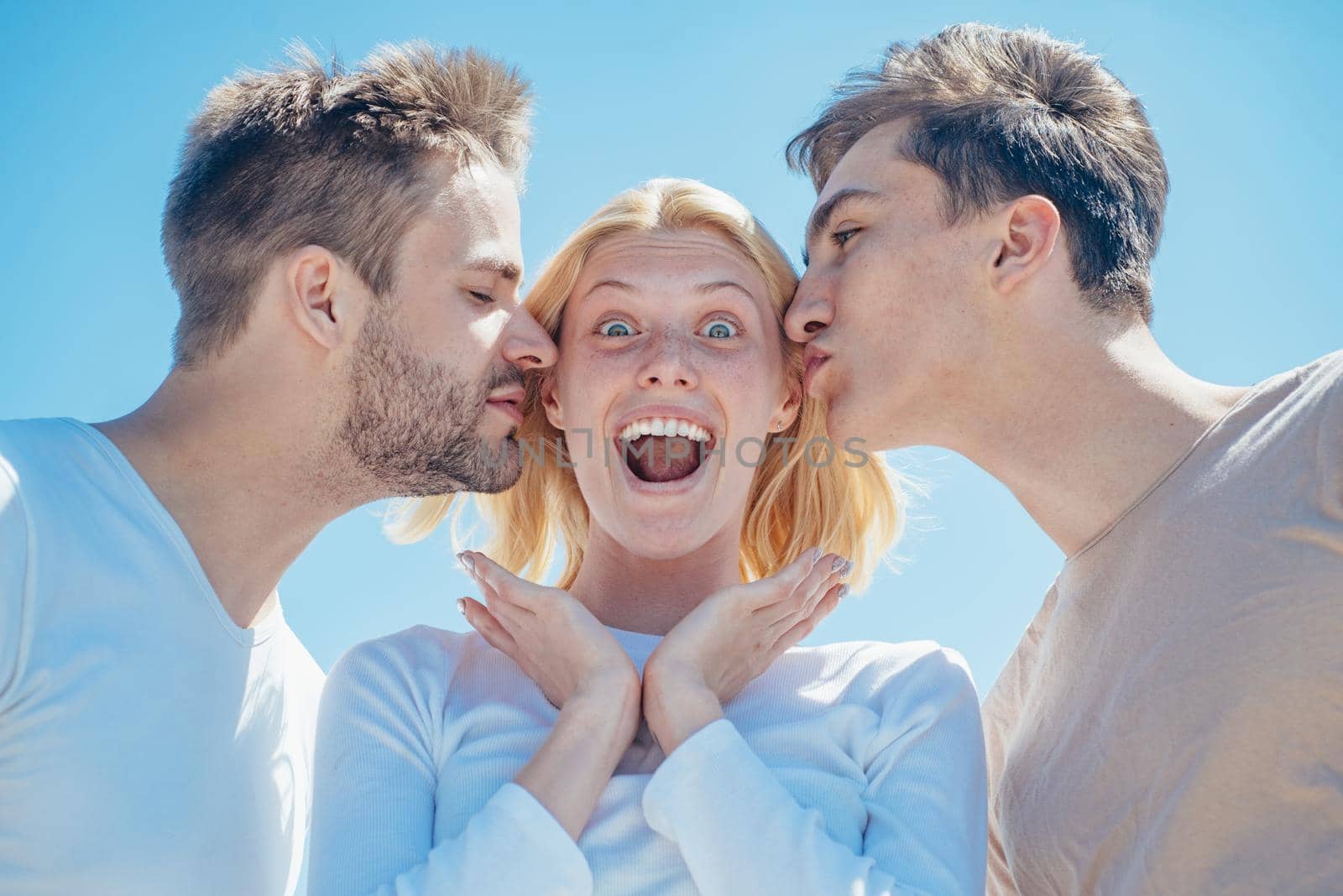 Blonde happy girl getting kisses from two handsome boys. Charming woman standing between two friends. Best friends concept. Group of friends at clear sky background. by Tverdokhlib