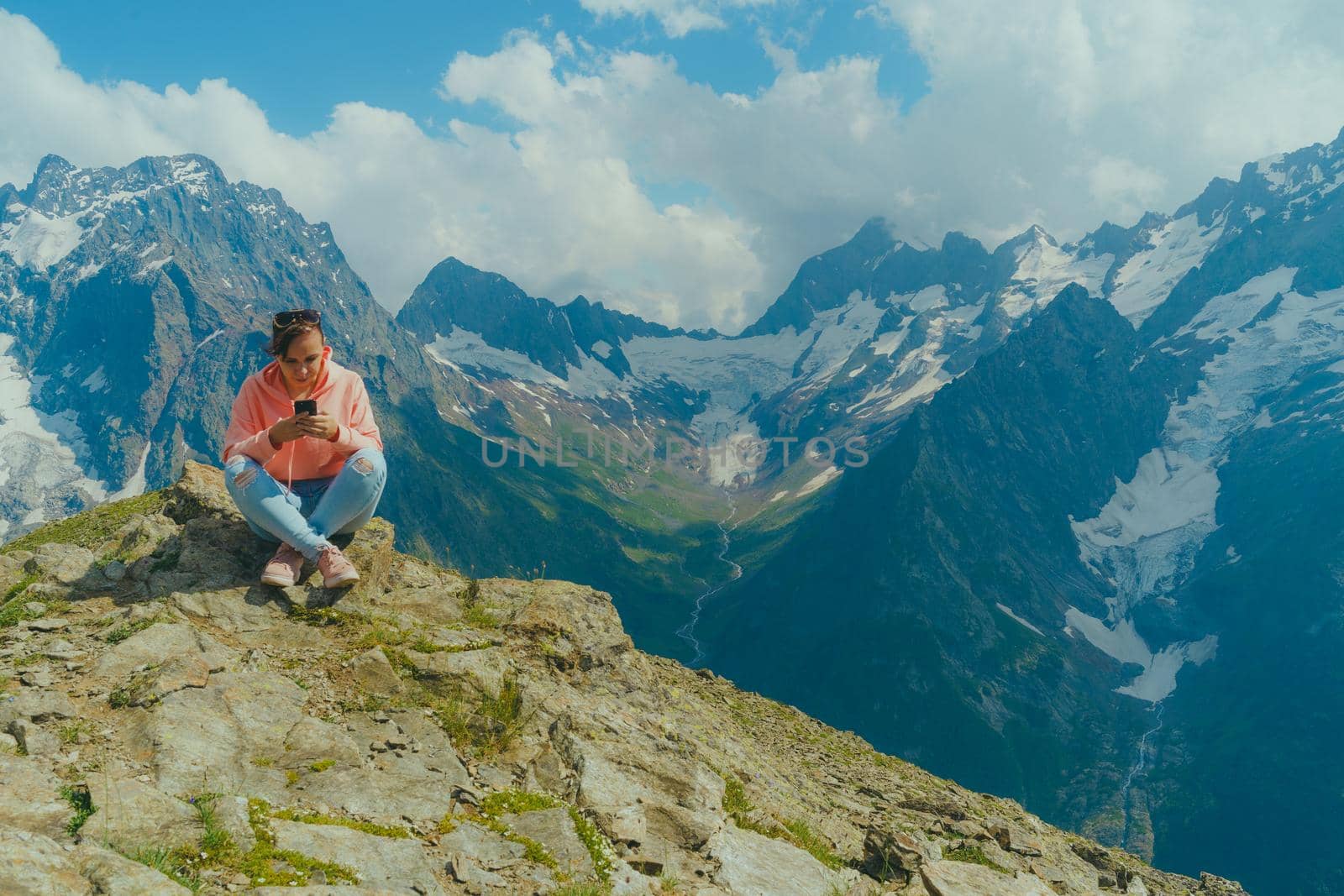Full body woman sitting on rock and browsing smartphone against cloudy sky on sunny day in mountains