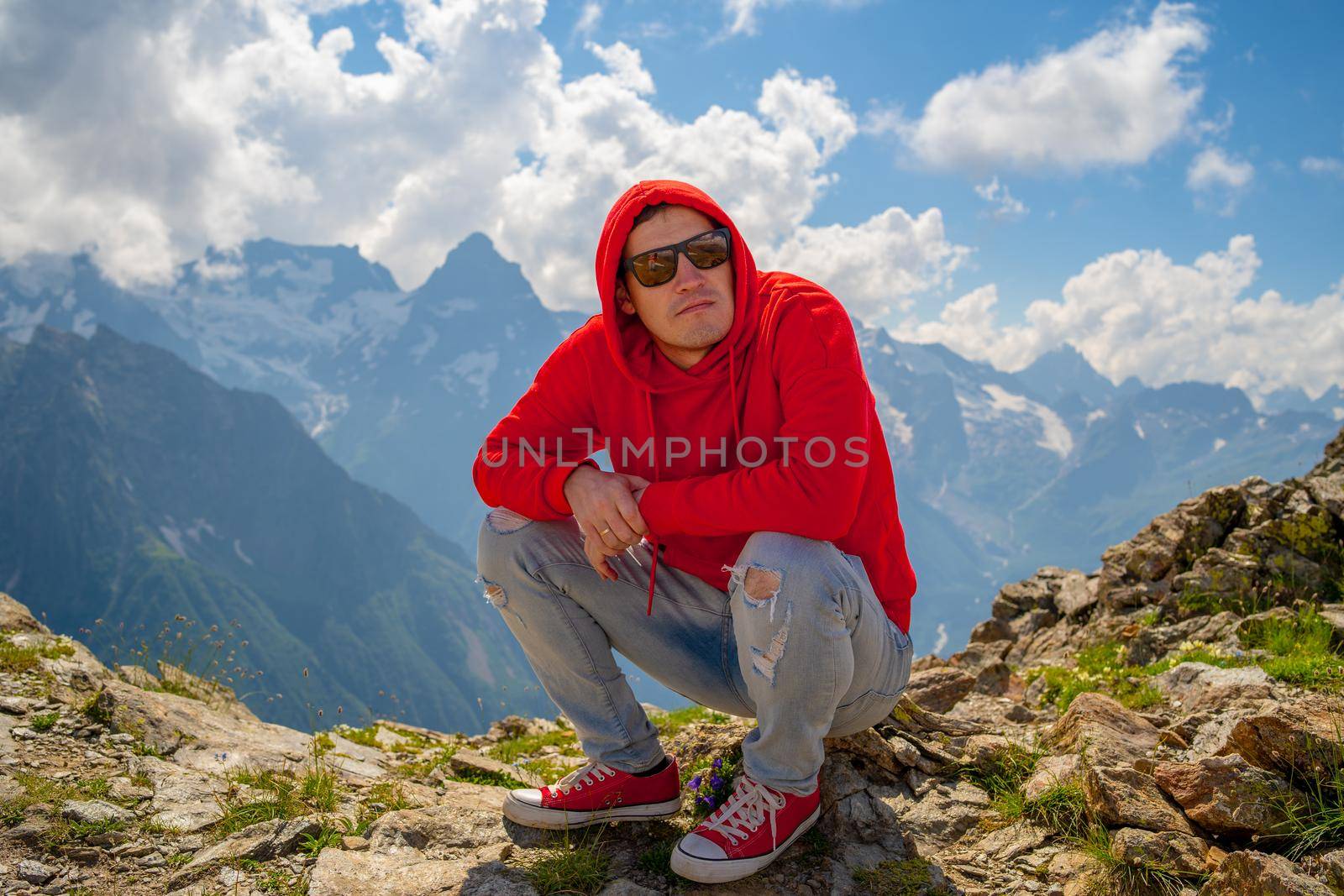 Young man in sunglasses sitting on mountain in sunny weather. Adult male in red hoodie with hood enjoying beautiful view in mountainous area. by epidemiks