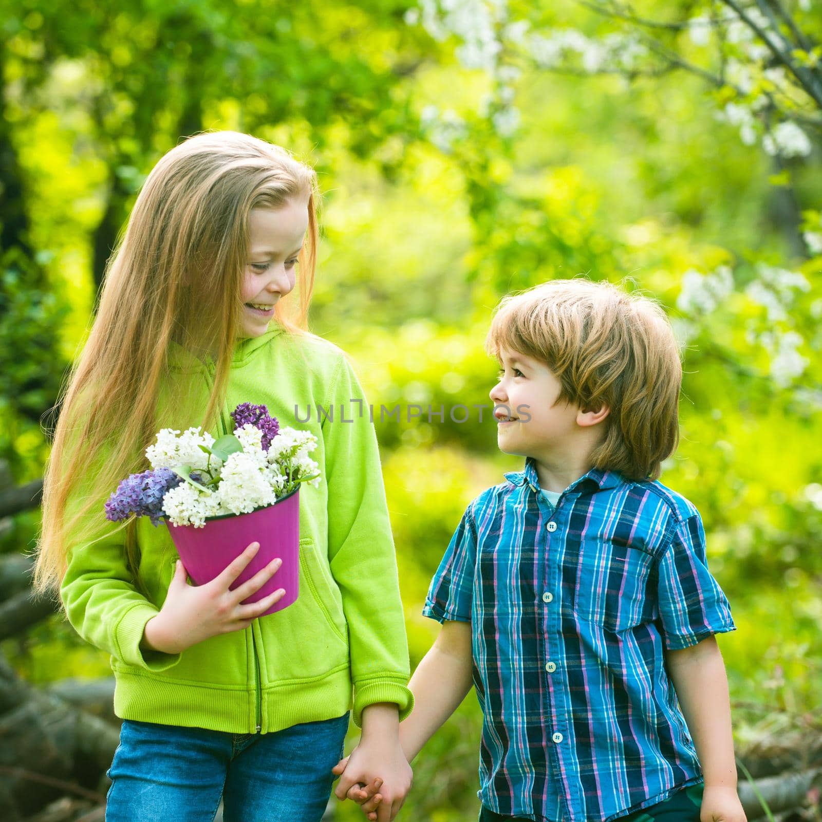 Cheerful smiling boy and girl look at each other and walk outdoor. First love and childhood concept. Little boy and girl holding hands. Children have fun in the spring garden. by Tverdokhlib