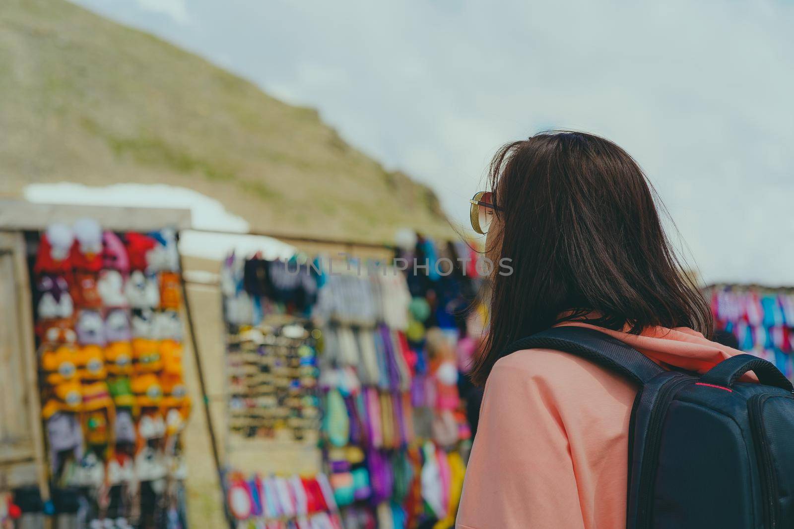 Female traveler on market in mountainous terrain. Woman tourist considering souvenirs in shopping booths. by epidemiks