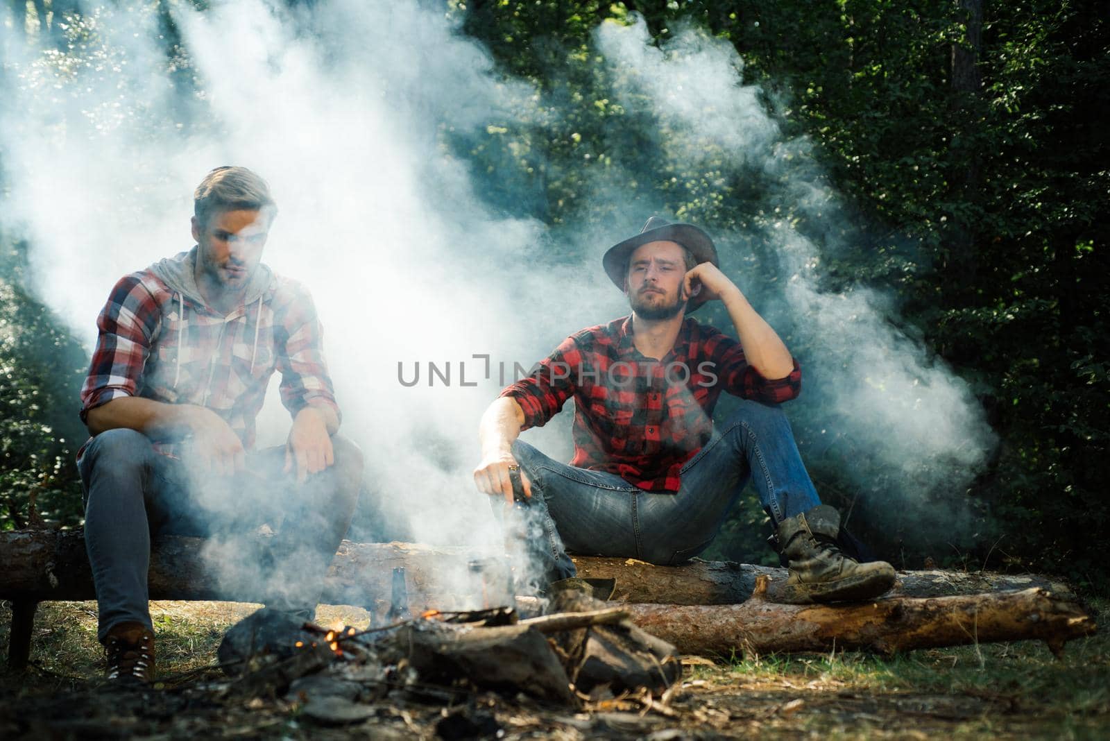 Good day for spring picnic in nature. Happy friends enjoying bonfire in nature. Friends having fun at camp fire. Two Happy people sitting around campfire with beer. Young guy having a picnic