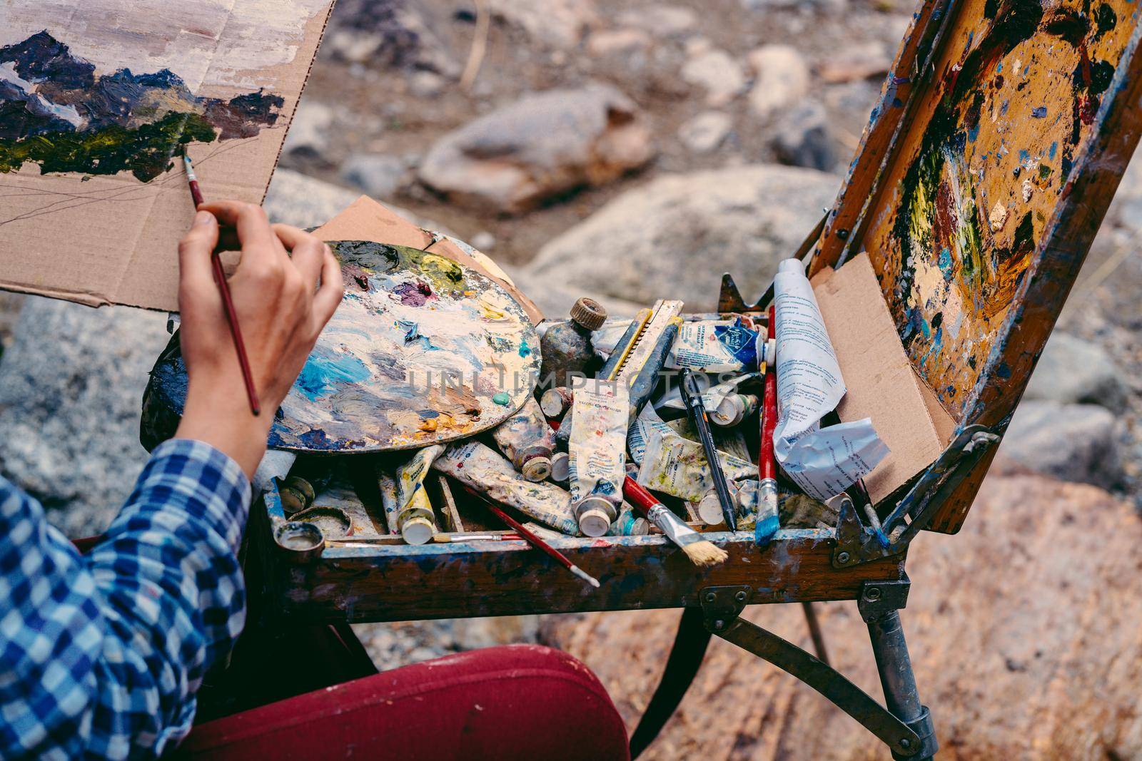 Close up of young woman drawing picture on cardboard on hike. Paintress depicting mountain landscape outdoors. by epidemiks