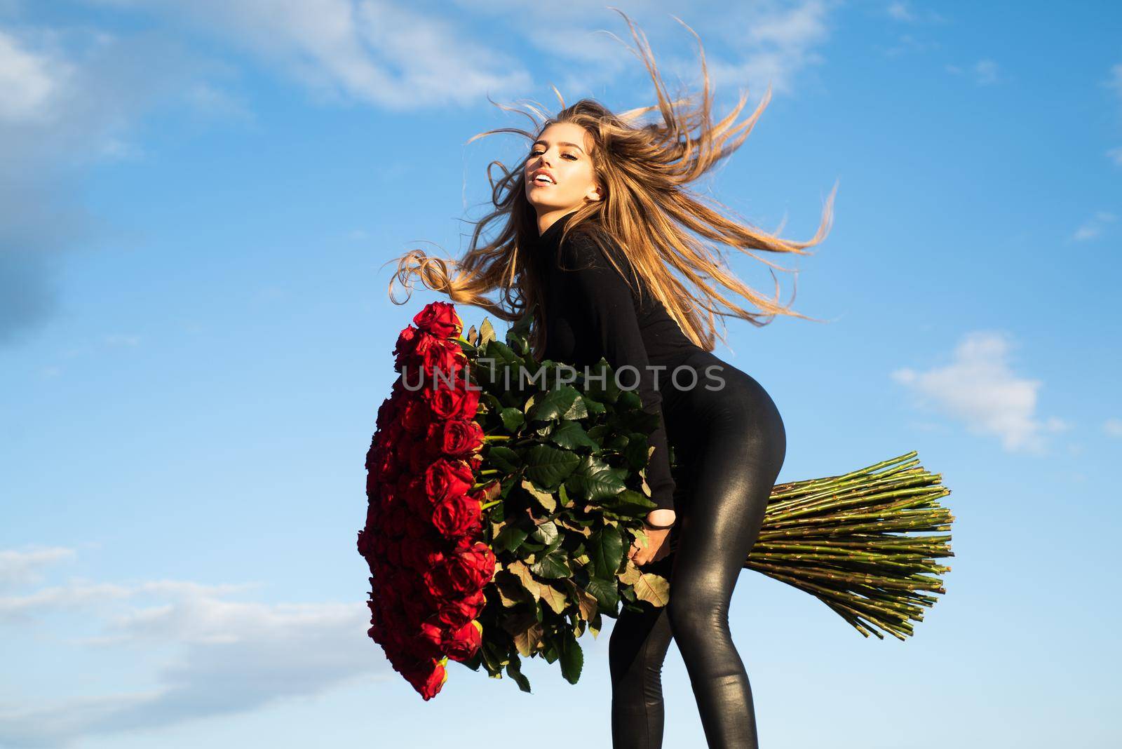 Beautiful girl on background sky. Girl and roses. Favorite flowers. Beautiful woman holding large bouquet of red roses. Gift with love. Happy girl hugs a bouquet