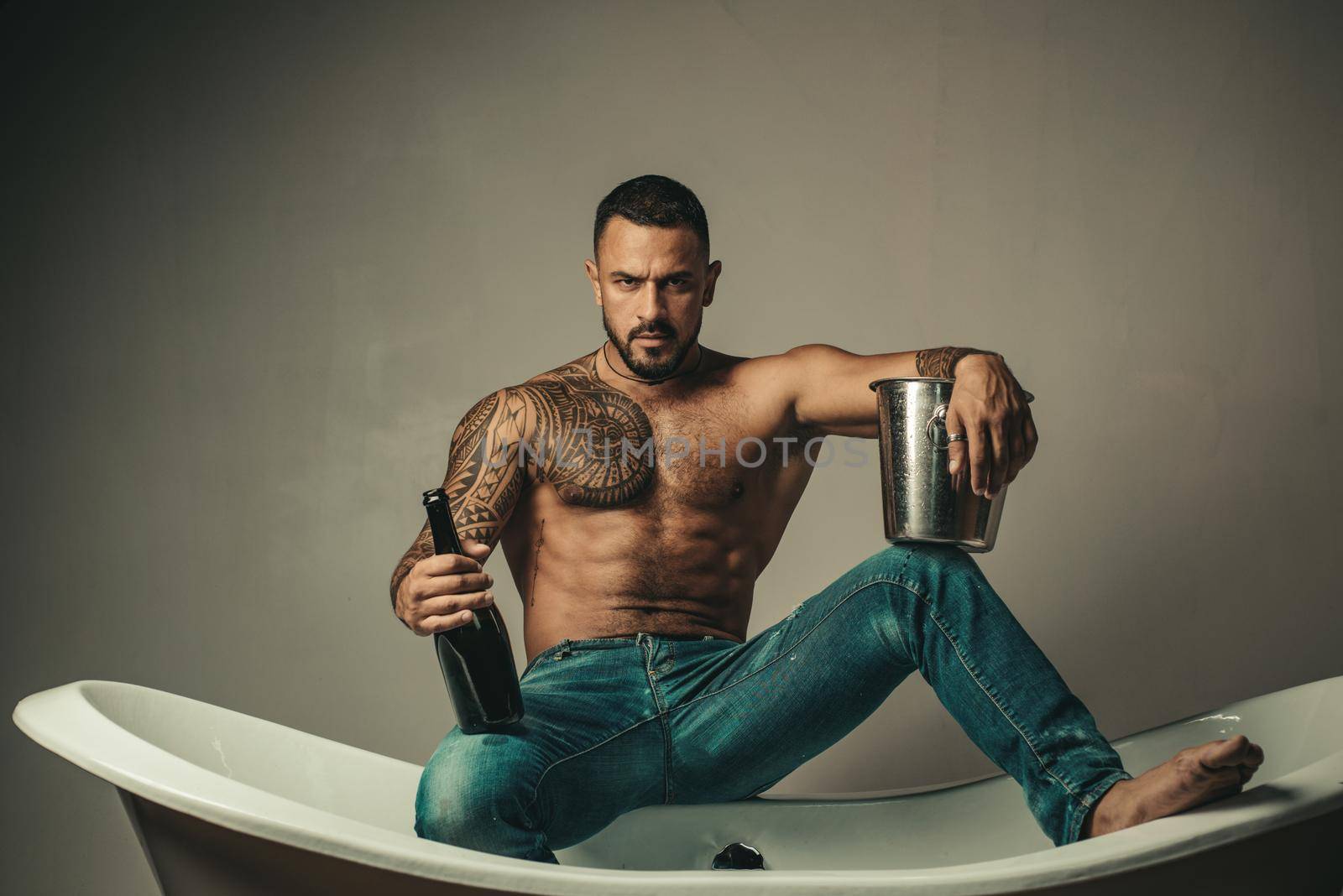 Handsome bearded shirtless man in jeans with sexy body in bathroom. Sexual macho man in bath. Strong muscular tattoed man holding champagne bottle and posing in bathroom