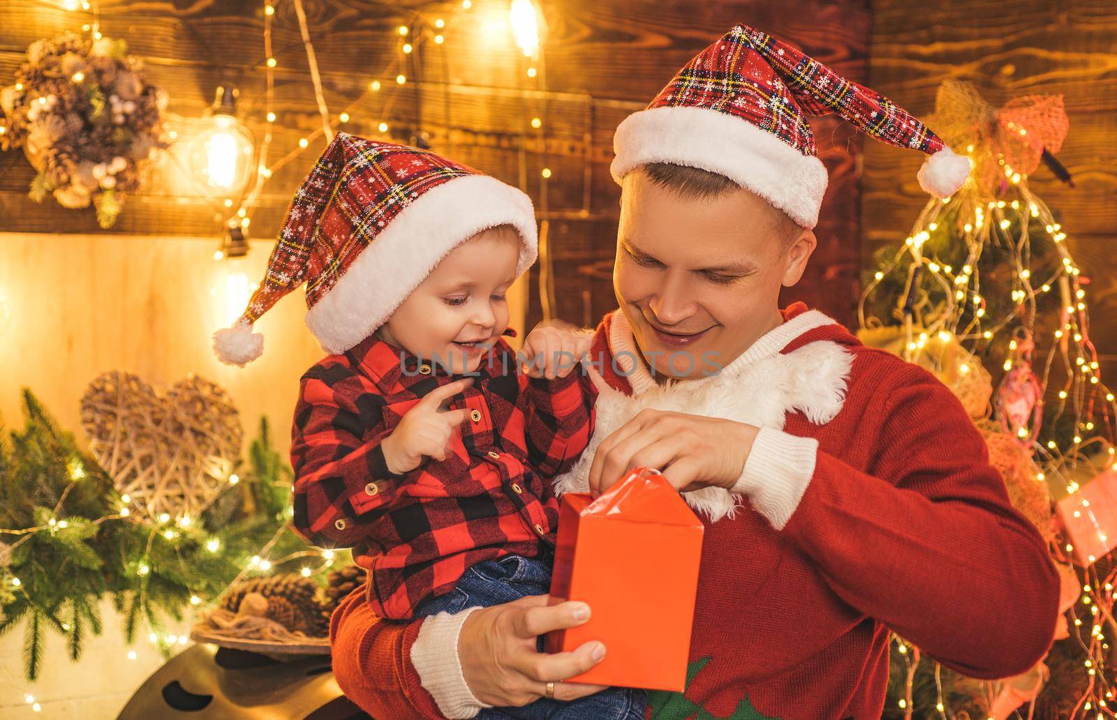 Smiling father and his son in Santa's hat open a Christmas gift present box. New Year tree and fire place at background. Christmas time. Childhood memories. Christmas child dreams