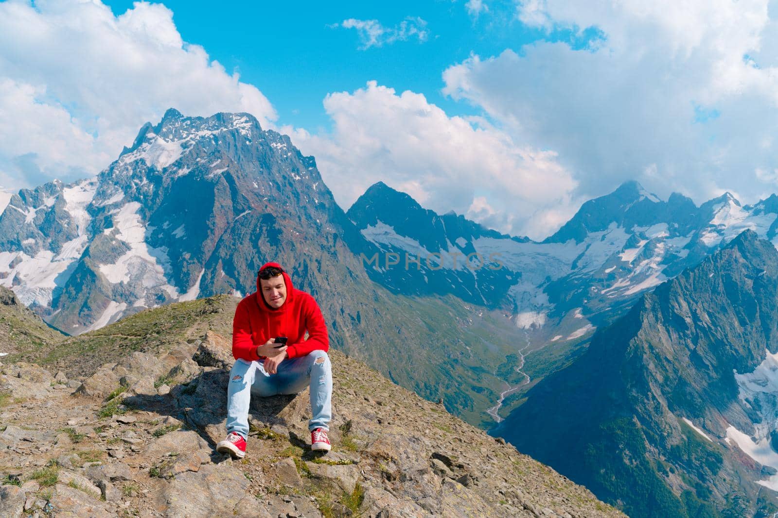 Man tourist sitting on rock with smartphone in mountains. Male traveler using mobile phone against cloudy sky on sunny day in mountainous terrain. by epidemiks