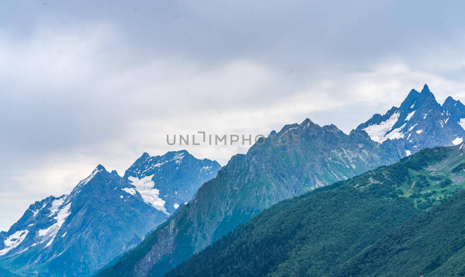 Beautiful mountain landscape in wooded area in summertime. Mighty mountains with snow and green array in cloudy weather. by epidemiks