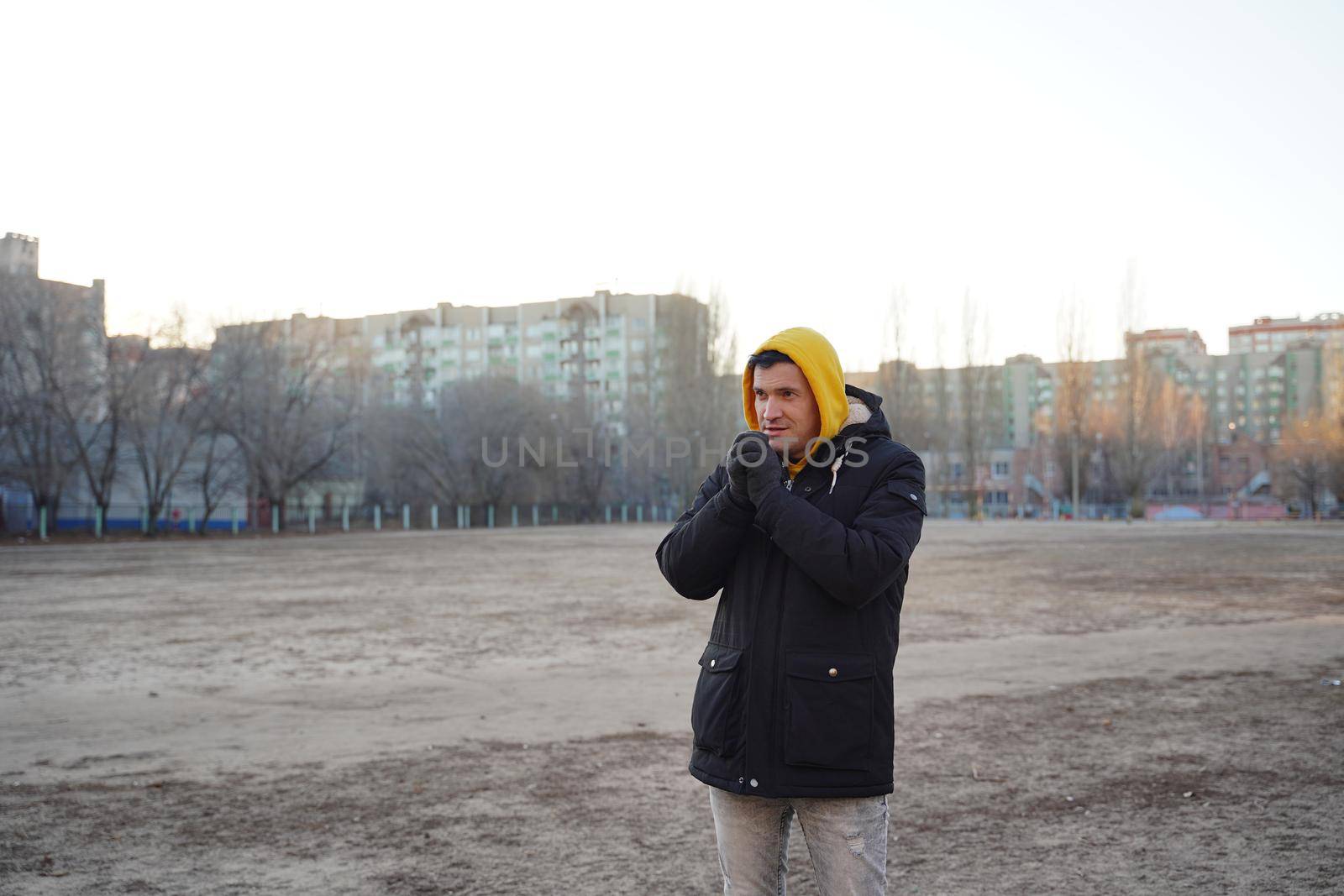 Young frozen man in yellow hood and black jacket. Adult male warming hands in gloves with his breath in cold weather. by epidemiks