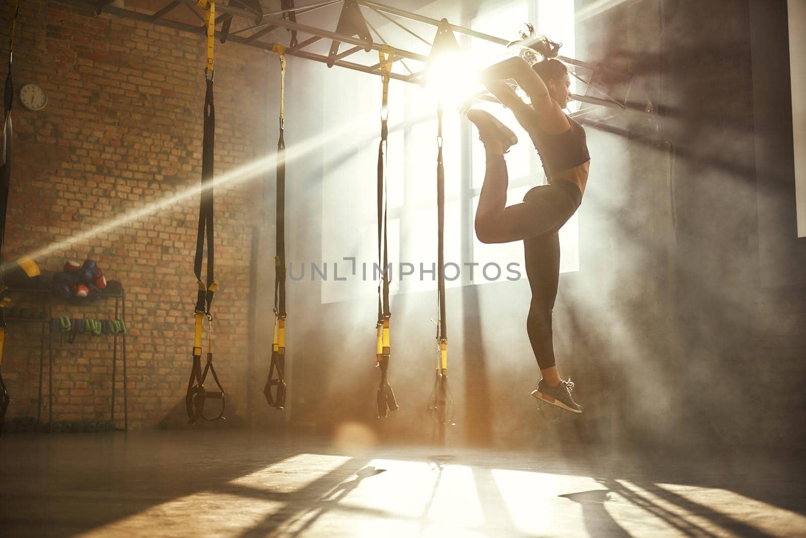 Perfect flexibility. Full length of athletic woman in black sport clothing jumping while exercising in professional semi-dark gym. by friendsstock