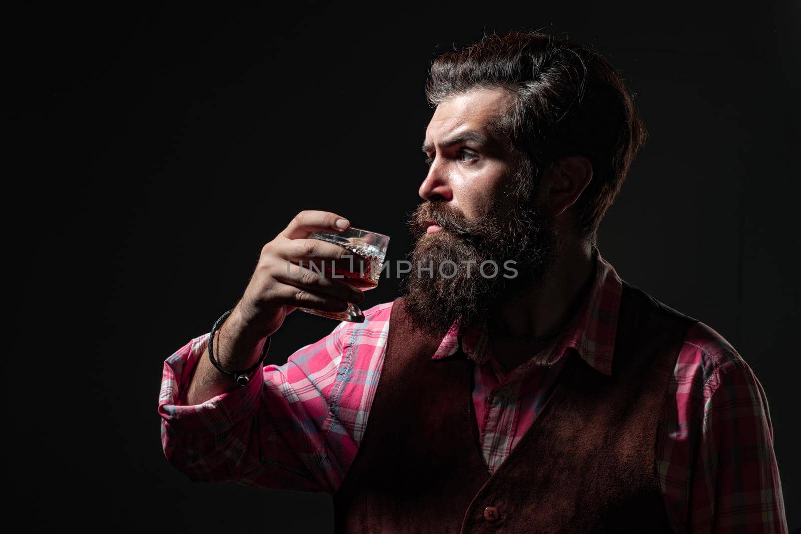 Handsome bearded businessman is drinking expensive whisky. Confident bearded man in black suit with glass of whisky in loft