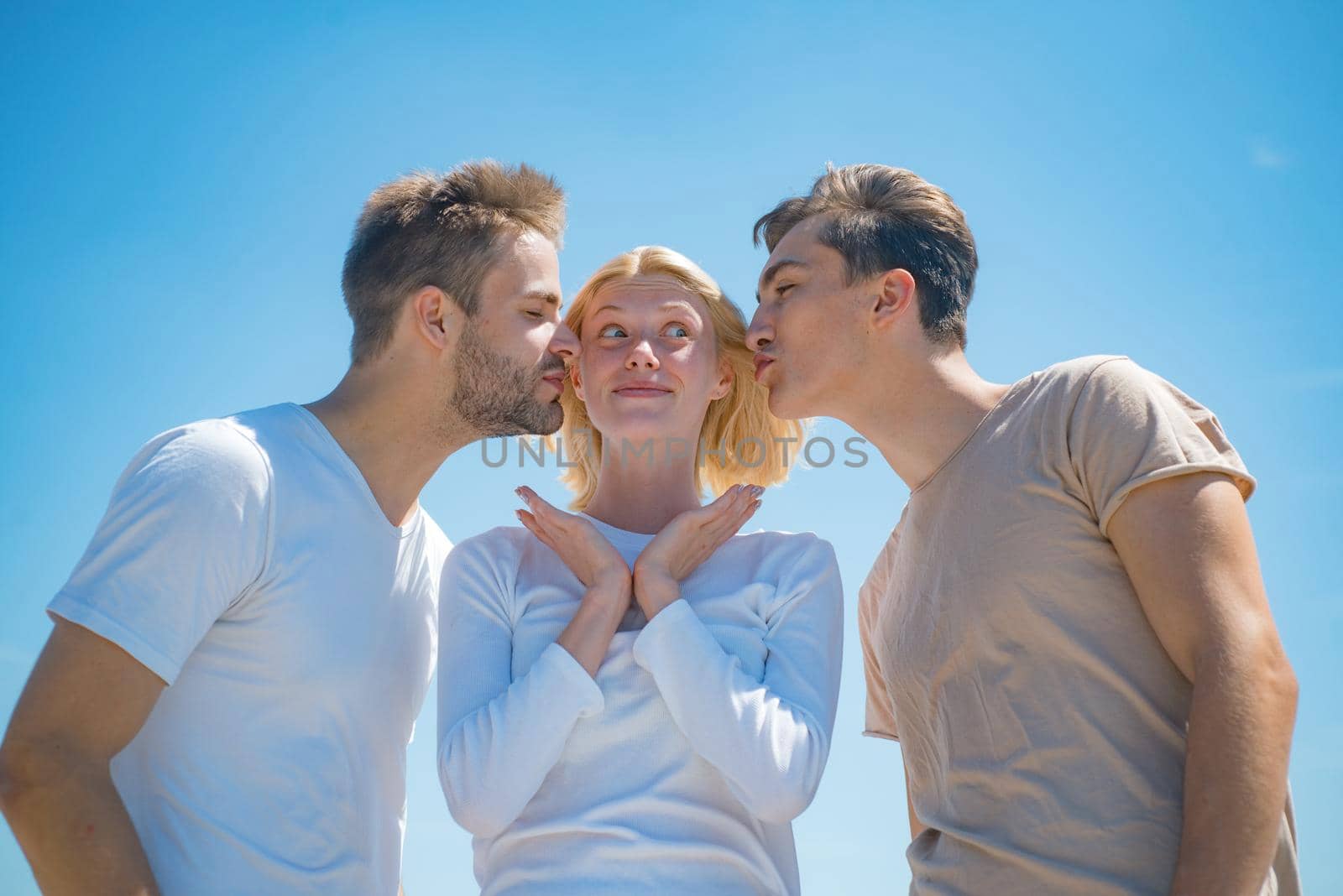 Company of happy young people spend leisure outdoors at sky background. Brothers kissing their surprised sister. Two boys and blonde woman at blue summer sky background