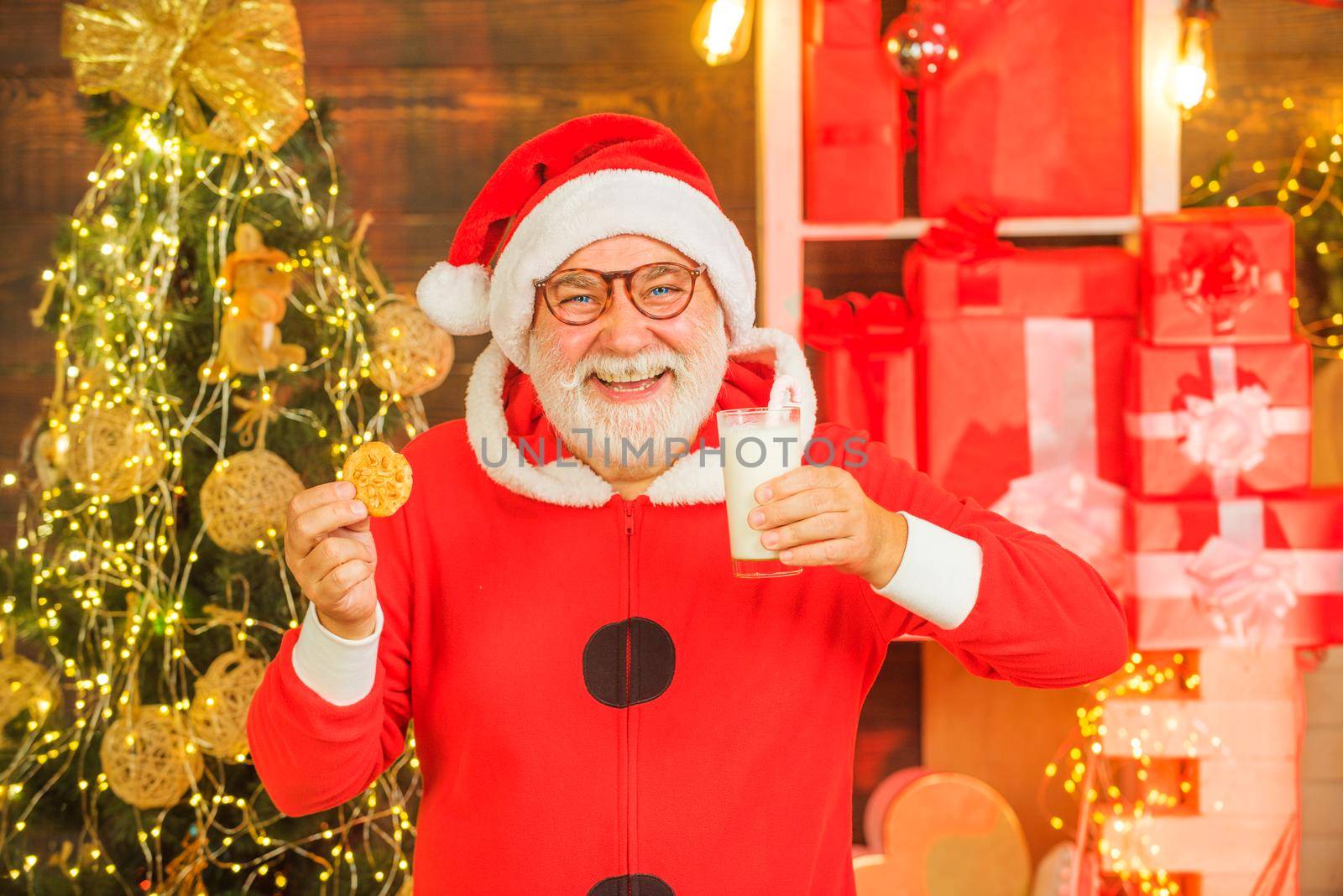 Santa Claus - bearded funny senior. Santa in home. Santa Claus eating cookies and drinking milk on Christmas Eve. Santa Claus enjoying in served gingerbread cake and milk