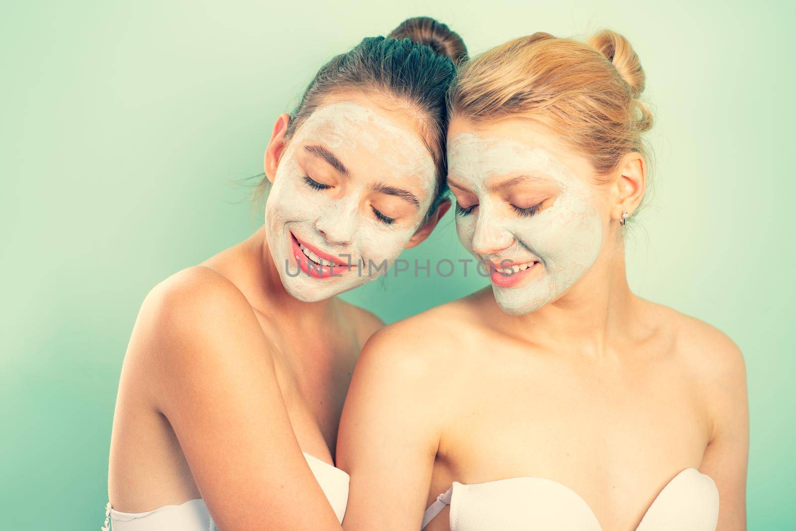Friendly young beautiful girls with bare shoulders apply organic face mask and smile. Blond and dark hair women make skincare. Friendship and young natural beauty concept