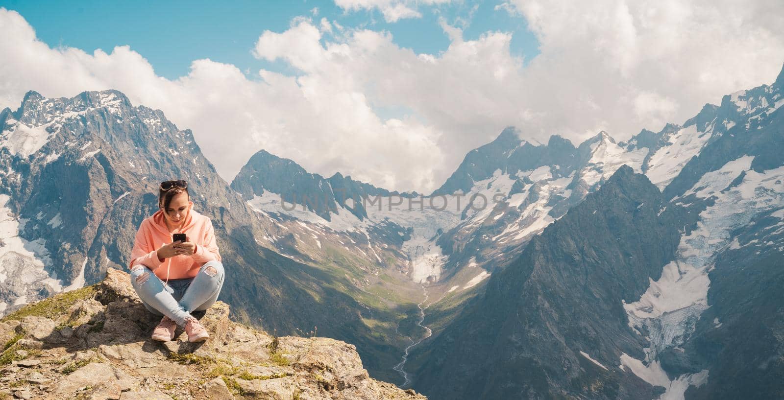 Female traveler using smartphone in mountains. Full body woman sitting on rock and browsing smartphone against cloudy sky on sunny day in mountains by epidemiks