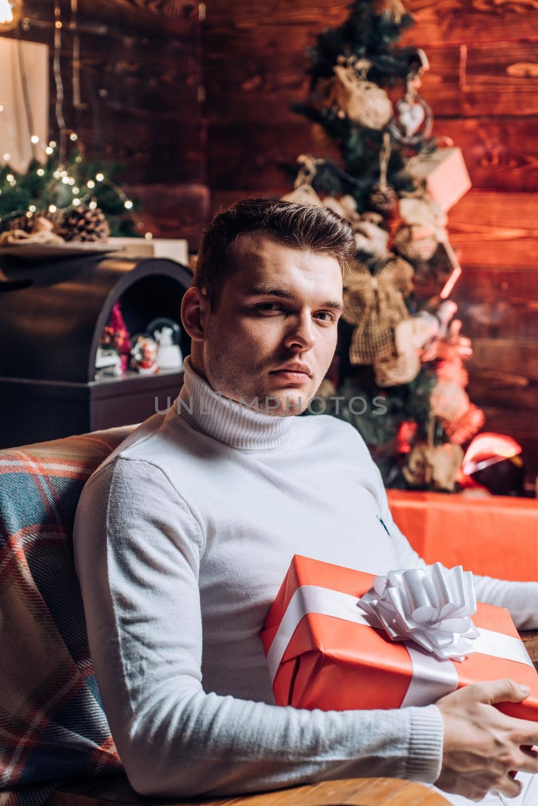 Handsome man holding christmas gift box and looking at camera at full decorated background. Warm cozy new year interior. Christmas eve. Happy new year