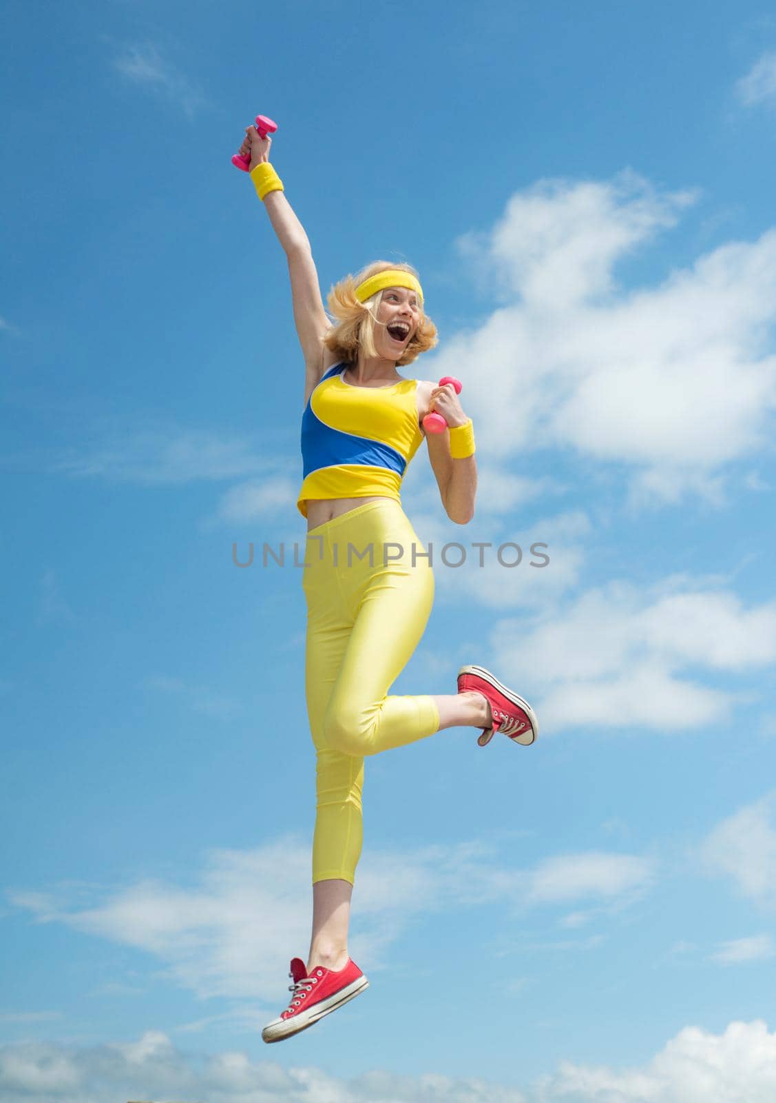 Young sporty excited girl jumping happily with hands up demonstrating exercises with pink dumbbells on sky background outdoor, panorama, copy space, sunrise light. Fashion and Trends in Sport. Health and fitness. Daily workout outdoors