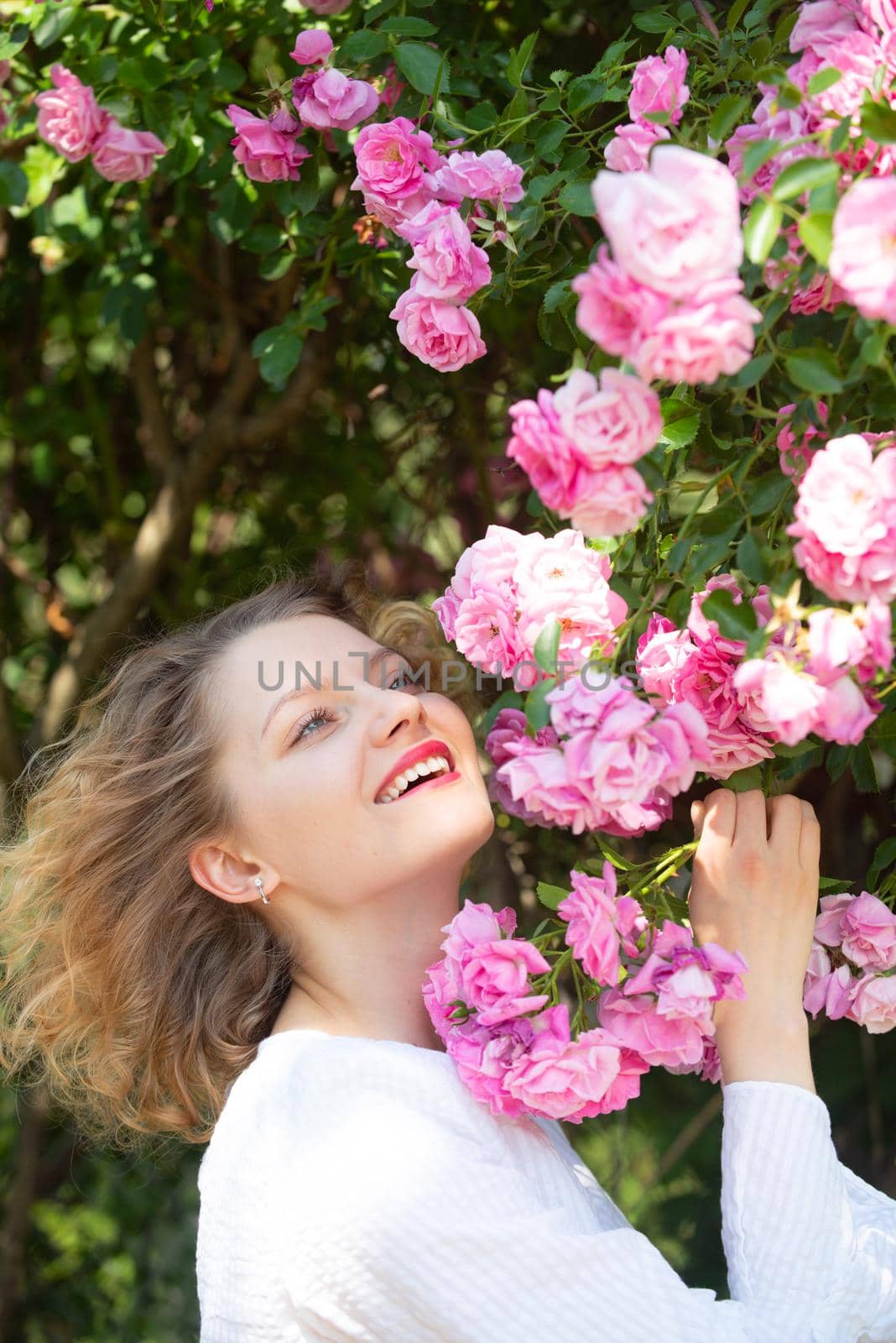 Charming girl at pastel pink rose blossom. Summer time. Beautiful young woman holding blooming rose flowers. Blossom around. Flowers of pink rose