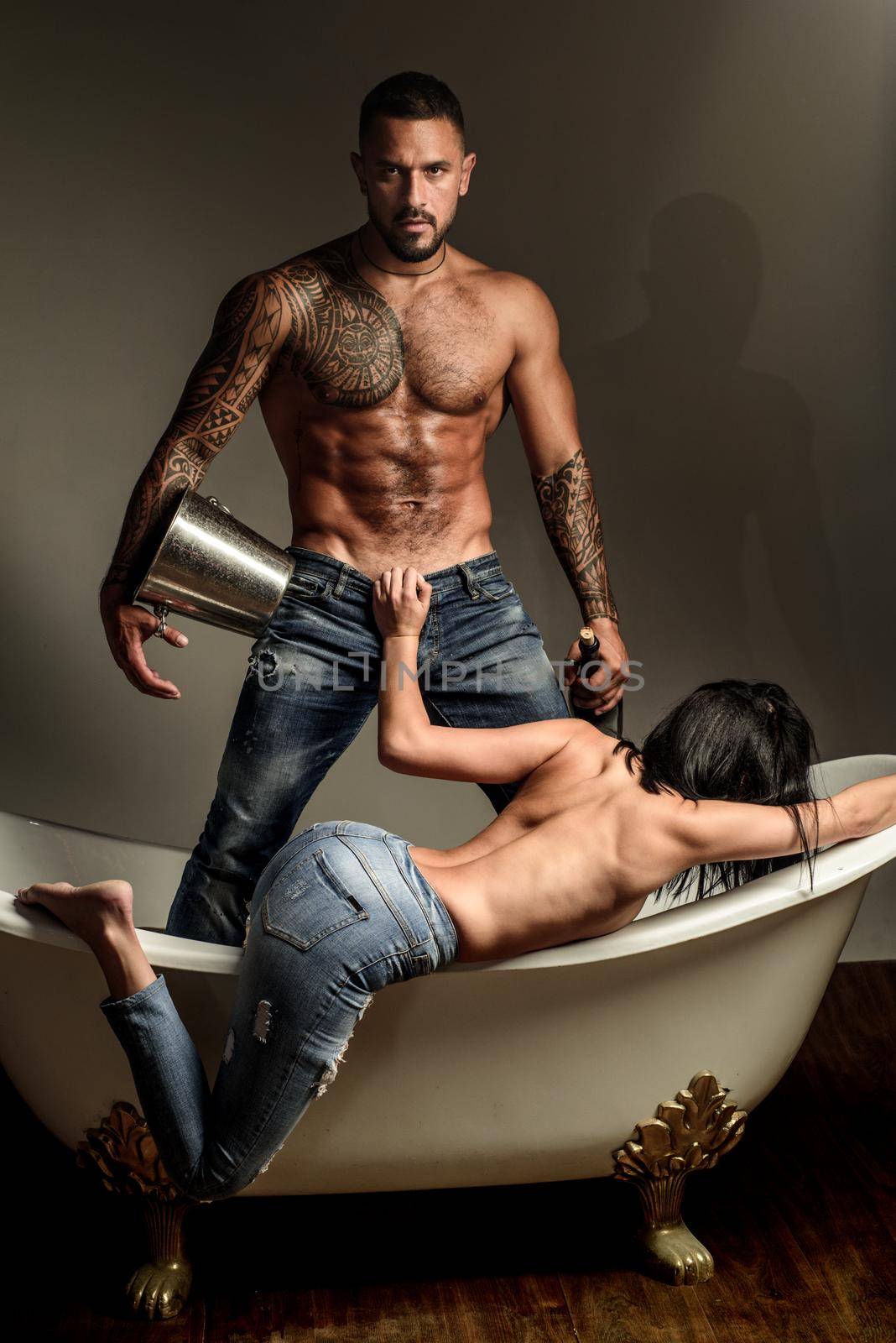 Brutal bearded tattooed man with sexy bare woman lying in bath. Handsome muscular man holds champagne cooler and bottle. Beautiful girl opens mans fly before sex. Couple foreplay