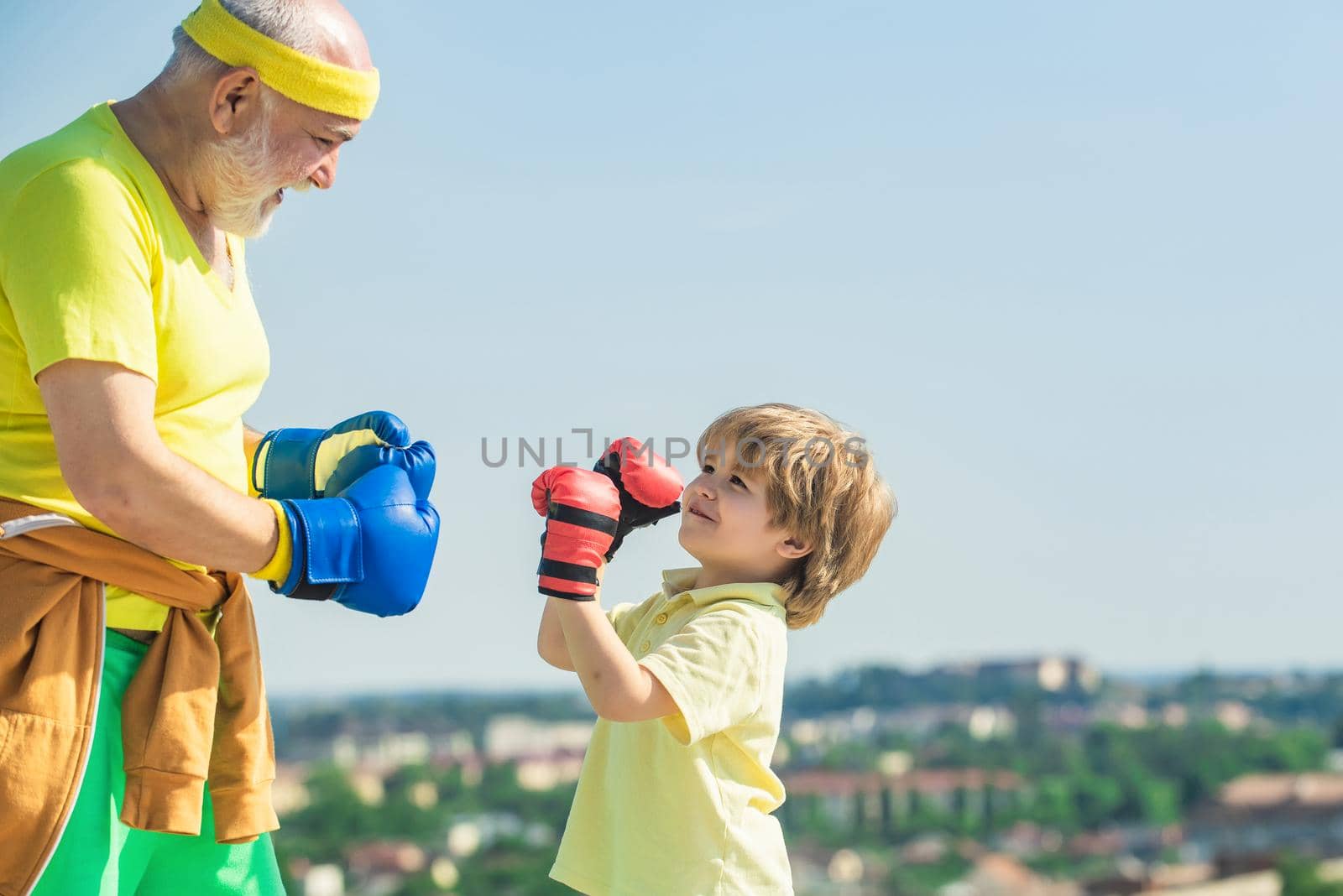 Grandpa and little child boy in boxing stance doing exercises with boxing gloves. Elderly man hitting punching bag. Grandfather and child fighting poses. by Tverdokhlib
