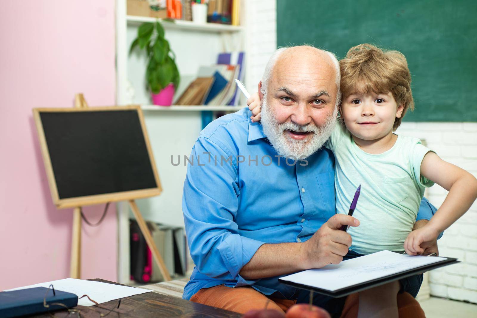 Happy cute Grandson and Grandfather sitting at a desk indoors. Funny little child having fun on blackboard background. A grandfather and a toddler are learning in class