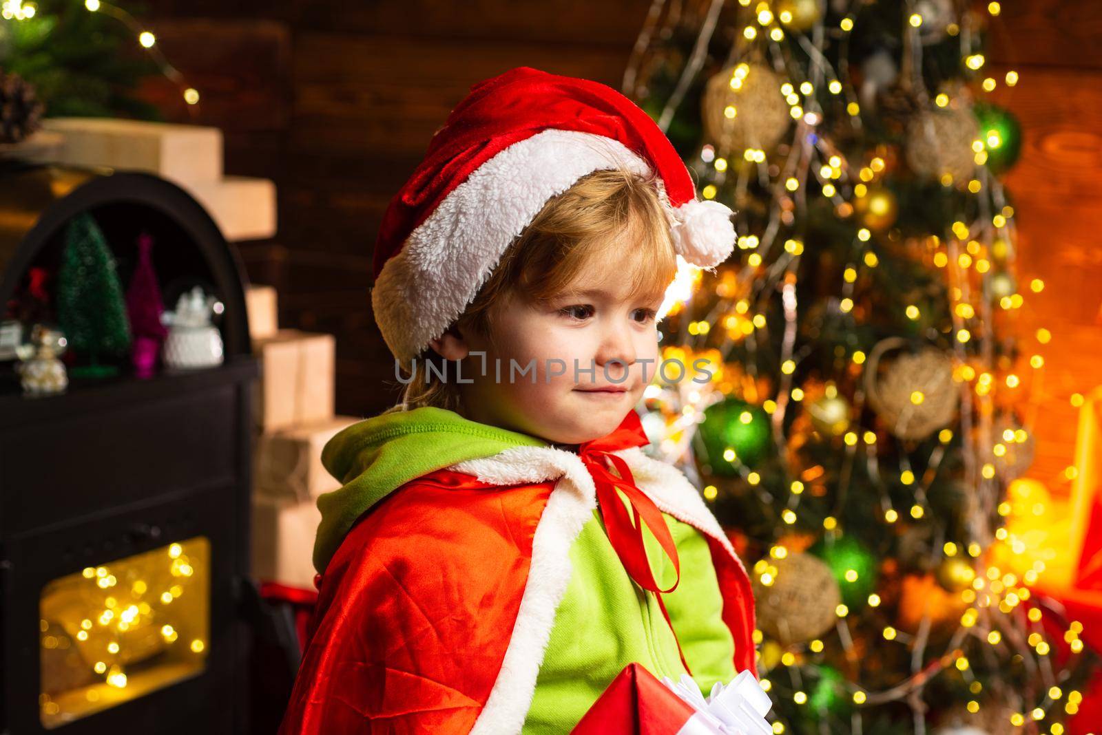 Happy little child in Santa's hat at Christmas tree background. Christmas time. Boy cute child cheerful mood play near christmas tree. Gifts for winter holidays near fire place. by Tverdokhlib