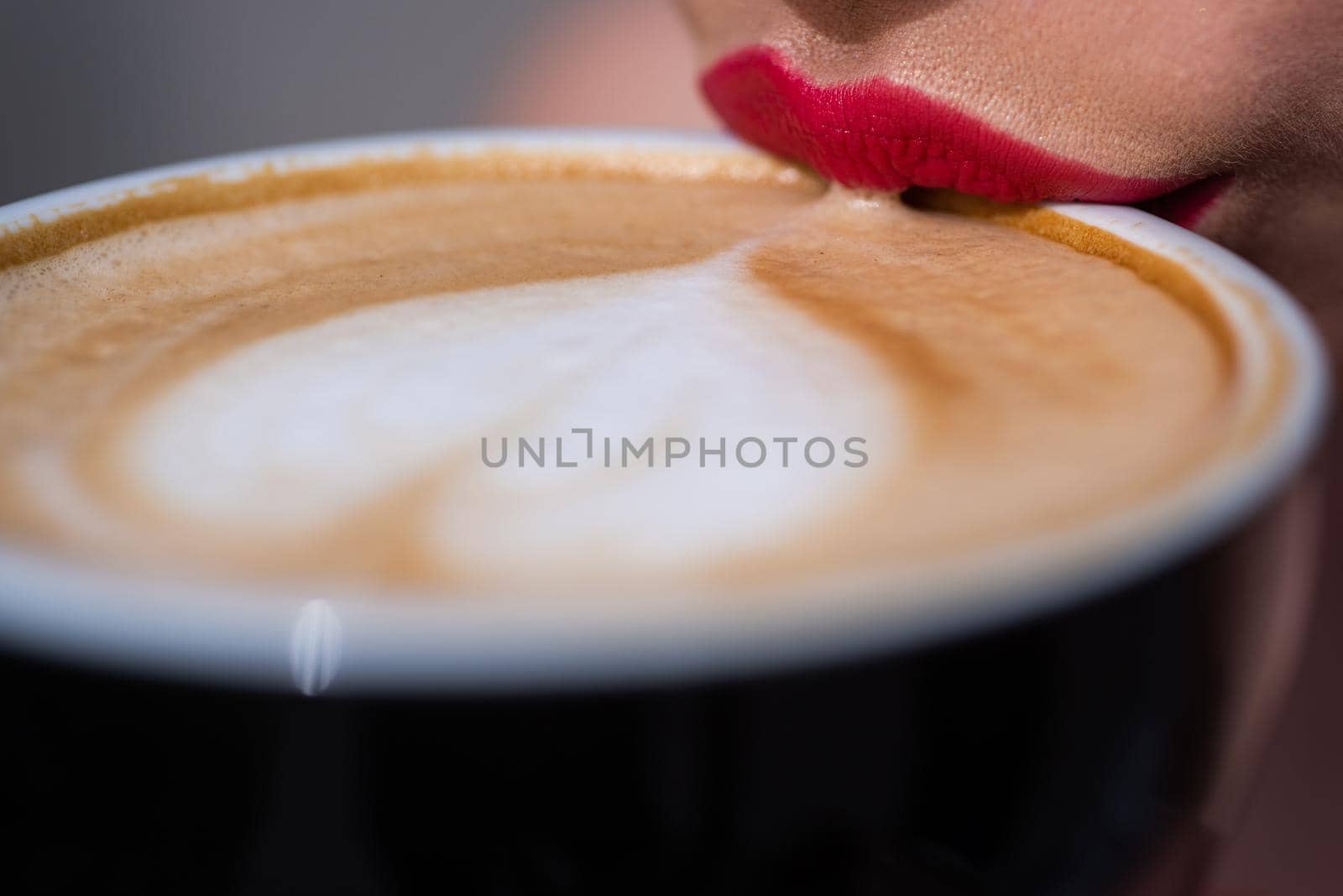Lips girl touch the cup with coffee. Coffee mug with heart shape on the froth. Closeup of cappuccino surface with heart shape and bright red female lips. Drinking
