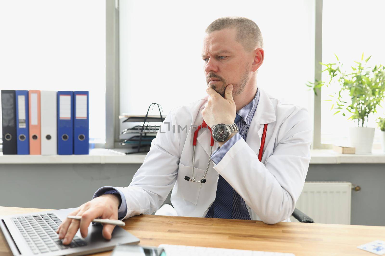 Pensive doctor looking at laptop screen and typing on keyboard in clinic by kuprevich