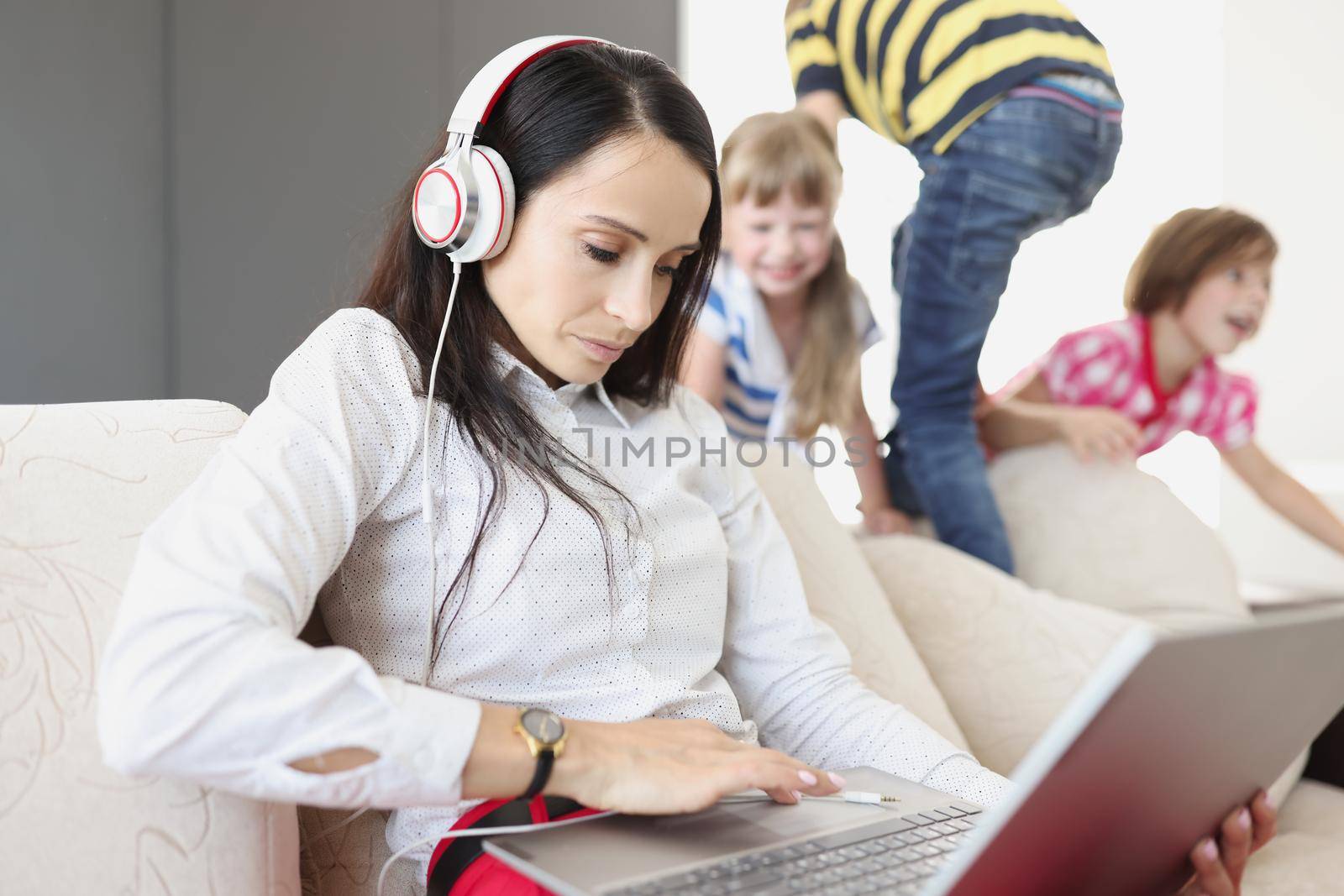 Young woman in headphones and with laptop sitting on sofa near playing children. Challenges of telecommuting during covid19 pandemic concept