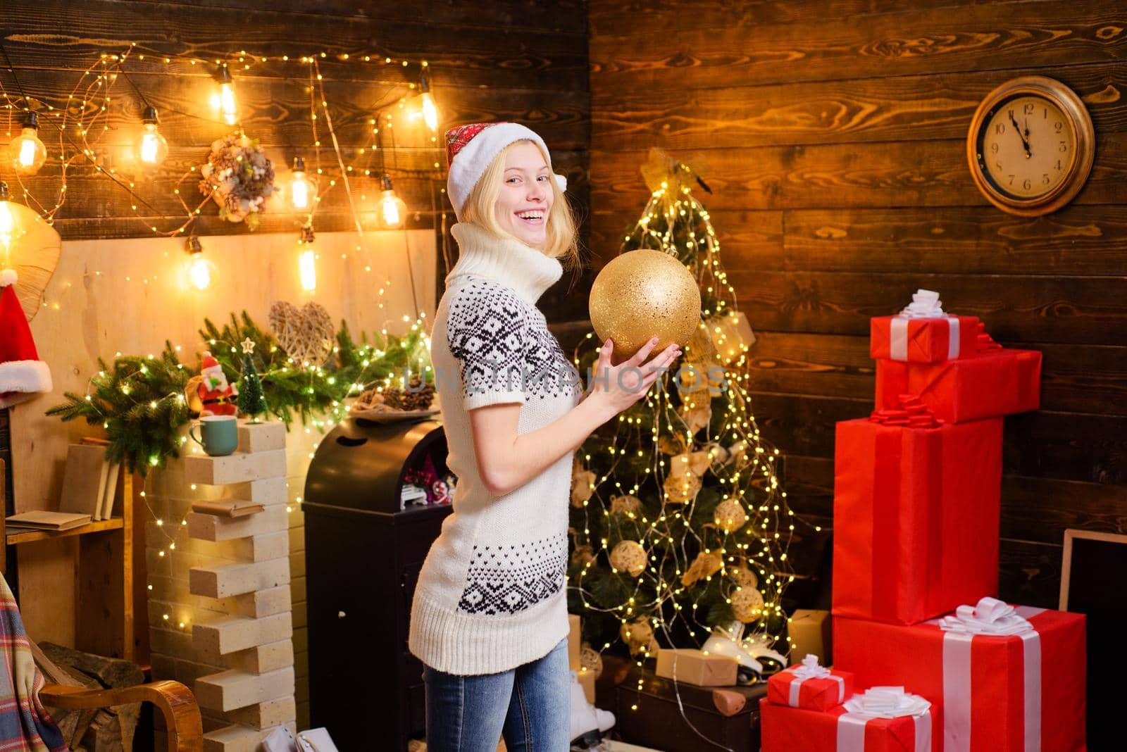 Happy smiling blonde girl wearing Santa's hat with fur and cozy warm sweater with deers and snowflackes. Snow Maiden beautiful girl hold a decorate ball at christmas background. by Tverdokhlib