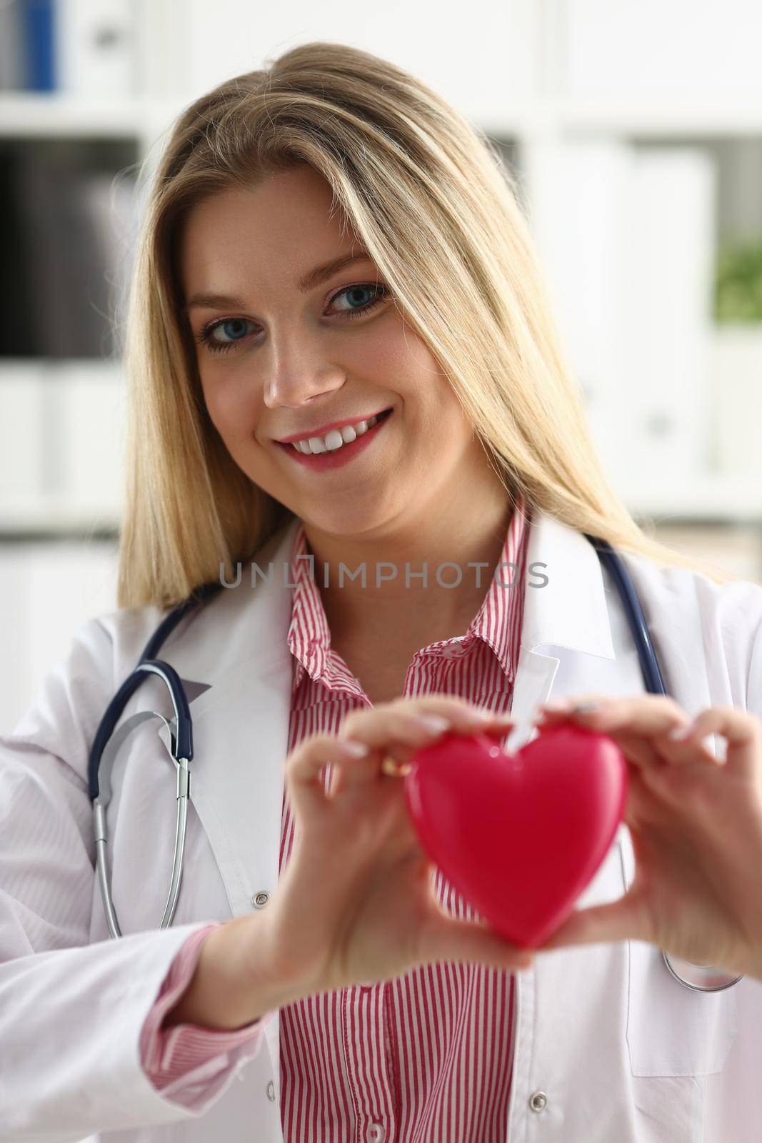 Beautiful smiling blond female doctor hold in arms red toy heart closeup. Cardio therapeutist student education CPR 911 life save physician make cardiac physical pulse rate measure arrhythmia