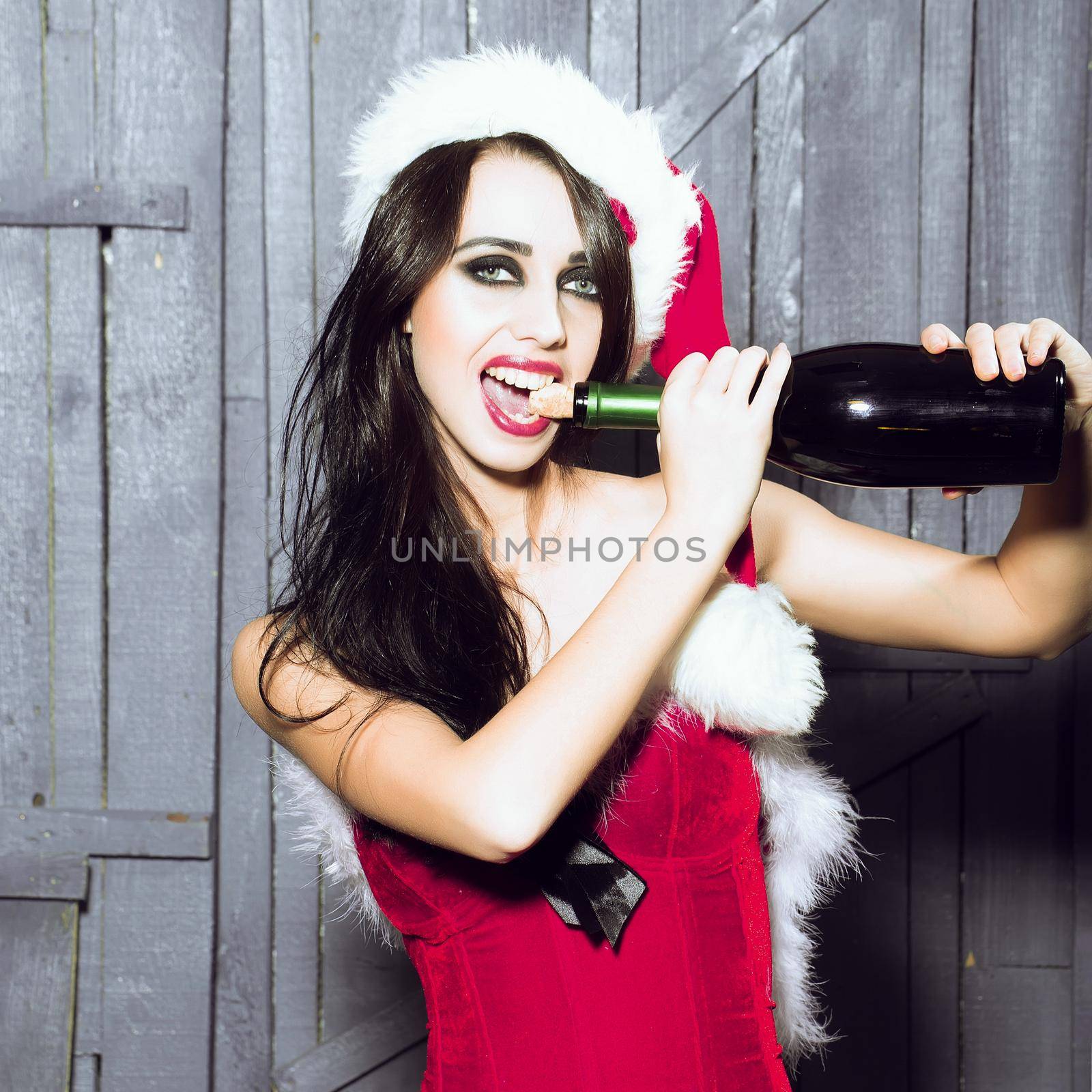 Woman pull out cork with teeth. Attractive woman in Santa Claus corset and red hat in studio on wooden background with corked wine bottle by Tverdokhlib