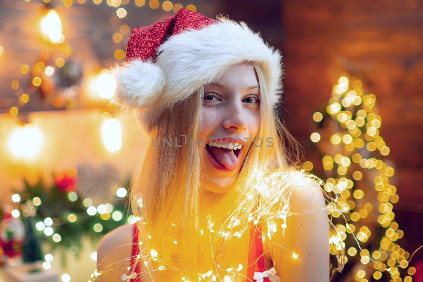 Beautiful young woman in Santa Claus clothes over christmas decorated interior background. Close up portrait of sexy girl flirting at camera