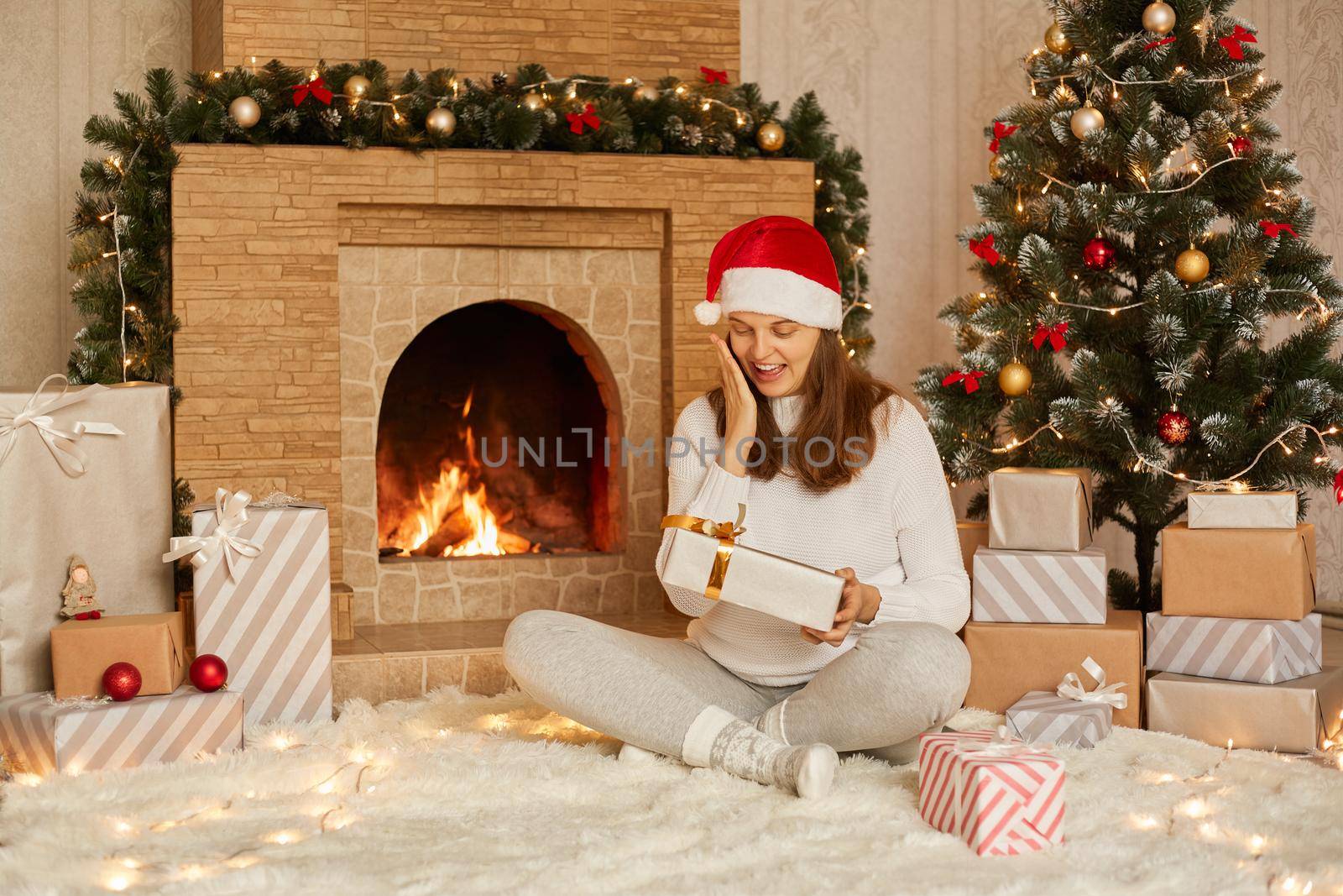 Beautiful young woman with dark hair, smiling, holding festive gift in hands while sitting near fireplace and Christmas tree. Happy girl dressed in santa hat, warm sweater and socks. by sementsovalesia