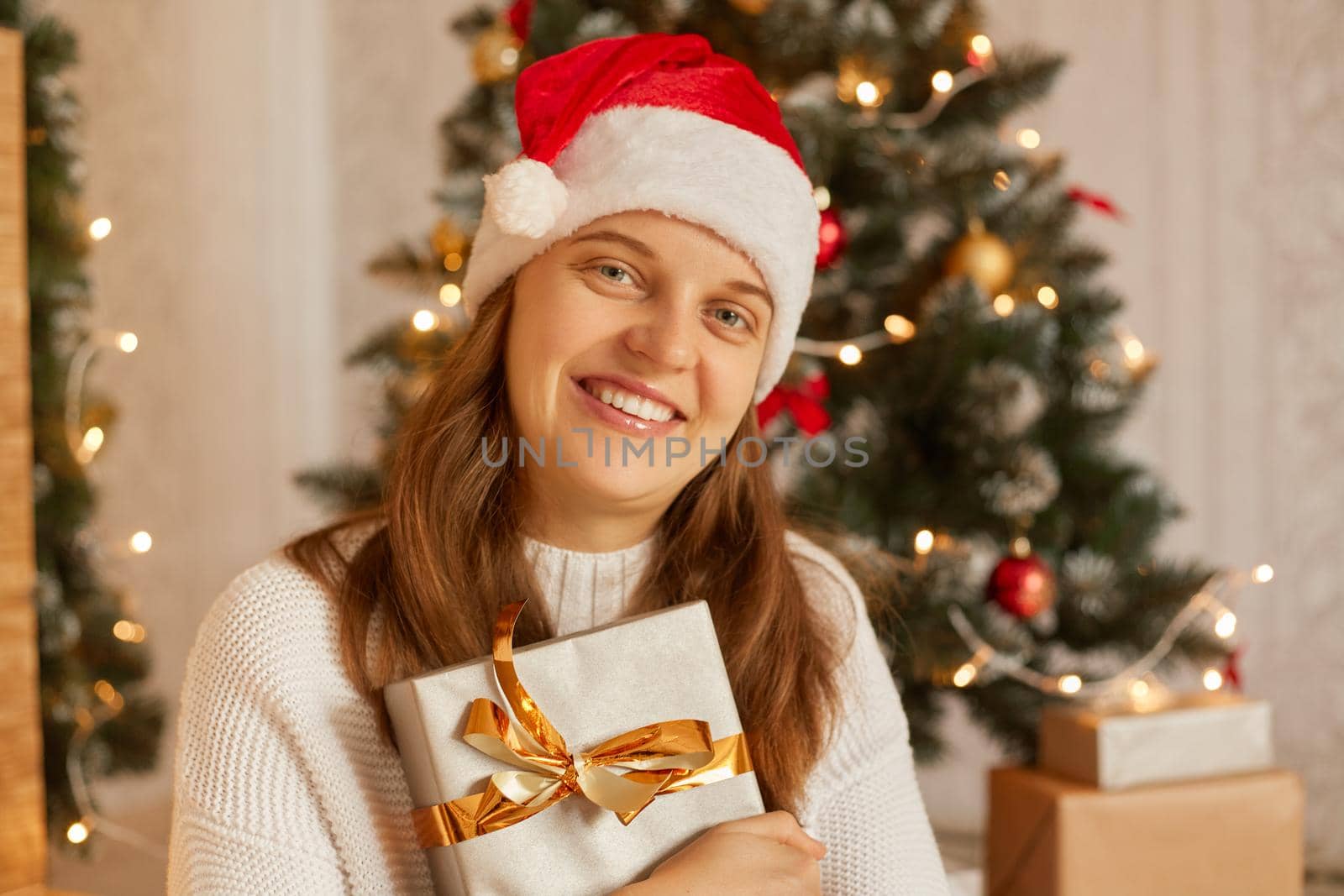 Portrait of smiling woman holding New Year gift in box with gold ribbon, happy female wearing santa hat and white sweater posing indoor with Christmas tree on background. by sementsovalesia