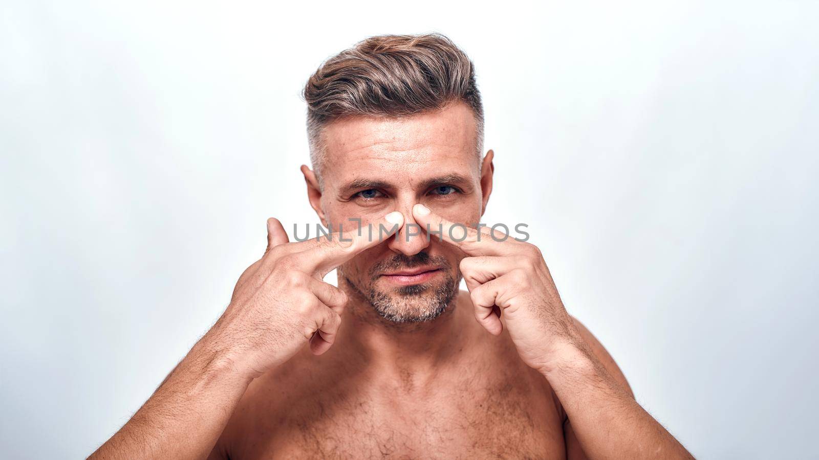 Everything should be perfect. Worried man with a stubble examining his nose and looking at camera while standing against grey background by friendsstock