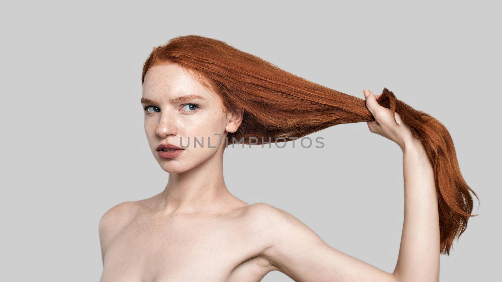 Studio shot of young and confident redhead woman holding her long silky hair while standing against grey background. Beauty concept. Hair care