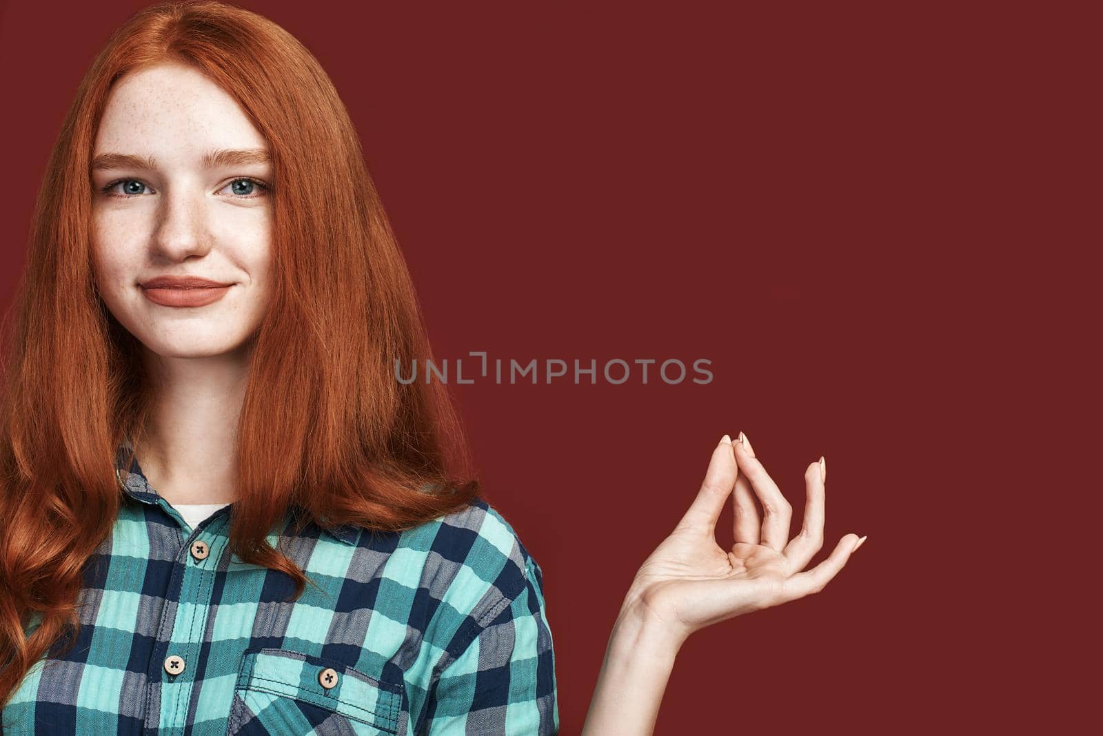 Photo of relaxed beautiful female in checkered shirt, making yoga gesture while looking at the camera, isolated over red studio background