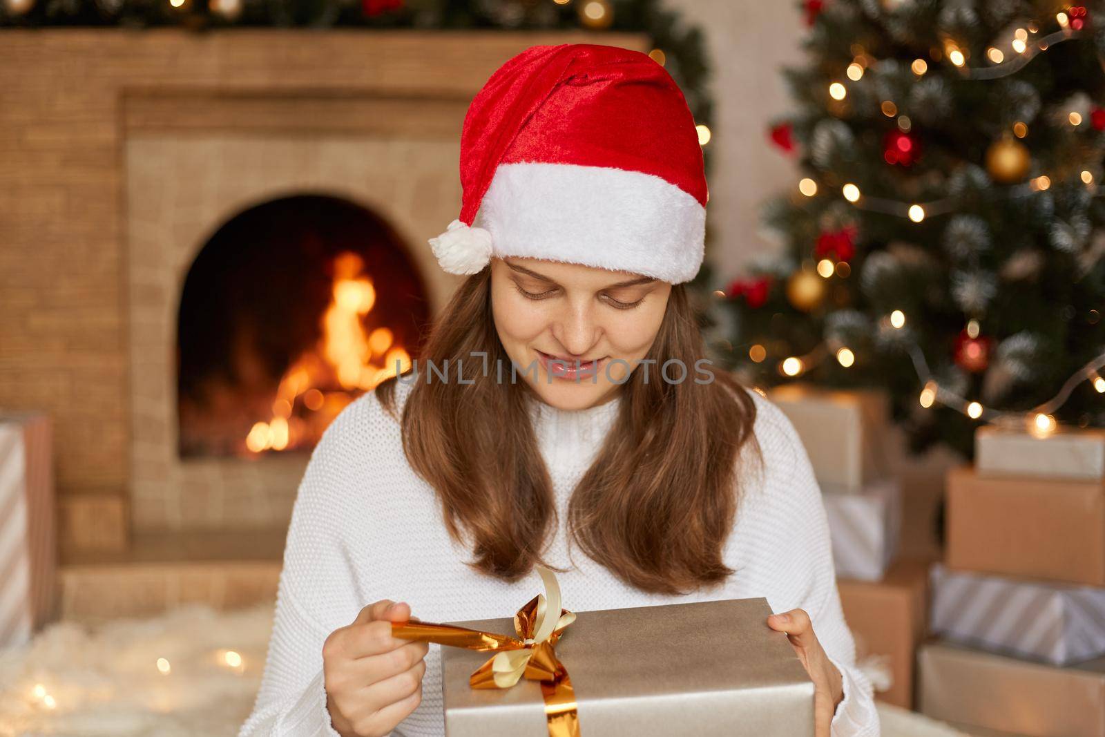 Adorable charming cheerful woman holds gift box and guess what inside, keeping fingers on ribbon and looking on box, sitting with fireplace and Christmas tree on background. by sementsovalesia
